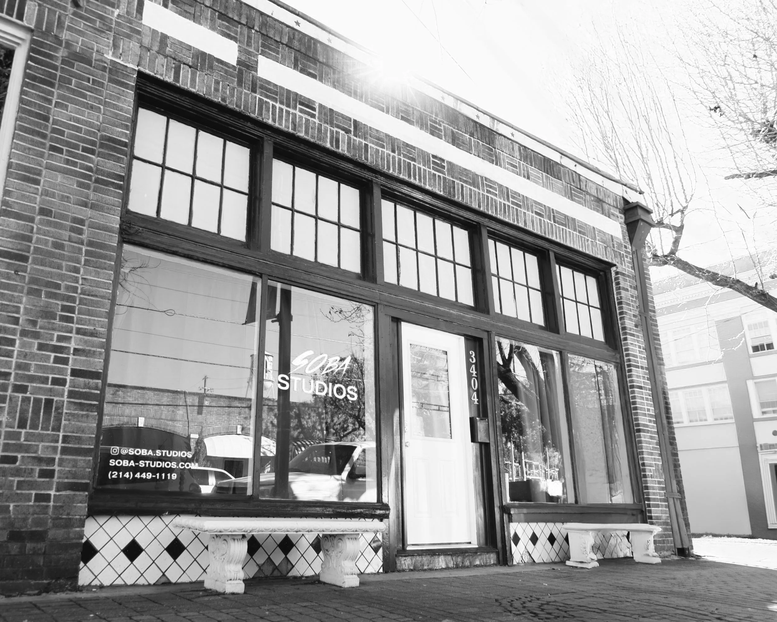 Black and white photo of a storefront with large windows and glass door, displaying 'SOBA STUDIOS' on the window. The building is made of brick with a decorative white band near the top. There are two white benches outside, and reflections of parked cars and trees can be seen in the window. The street appears quiet and sunny.