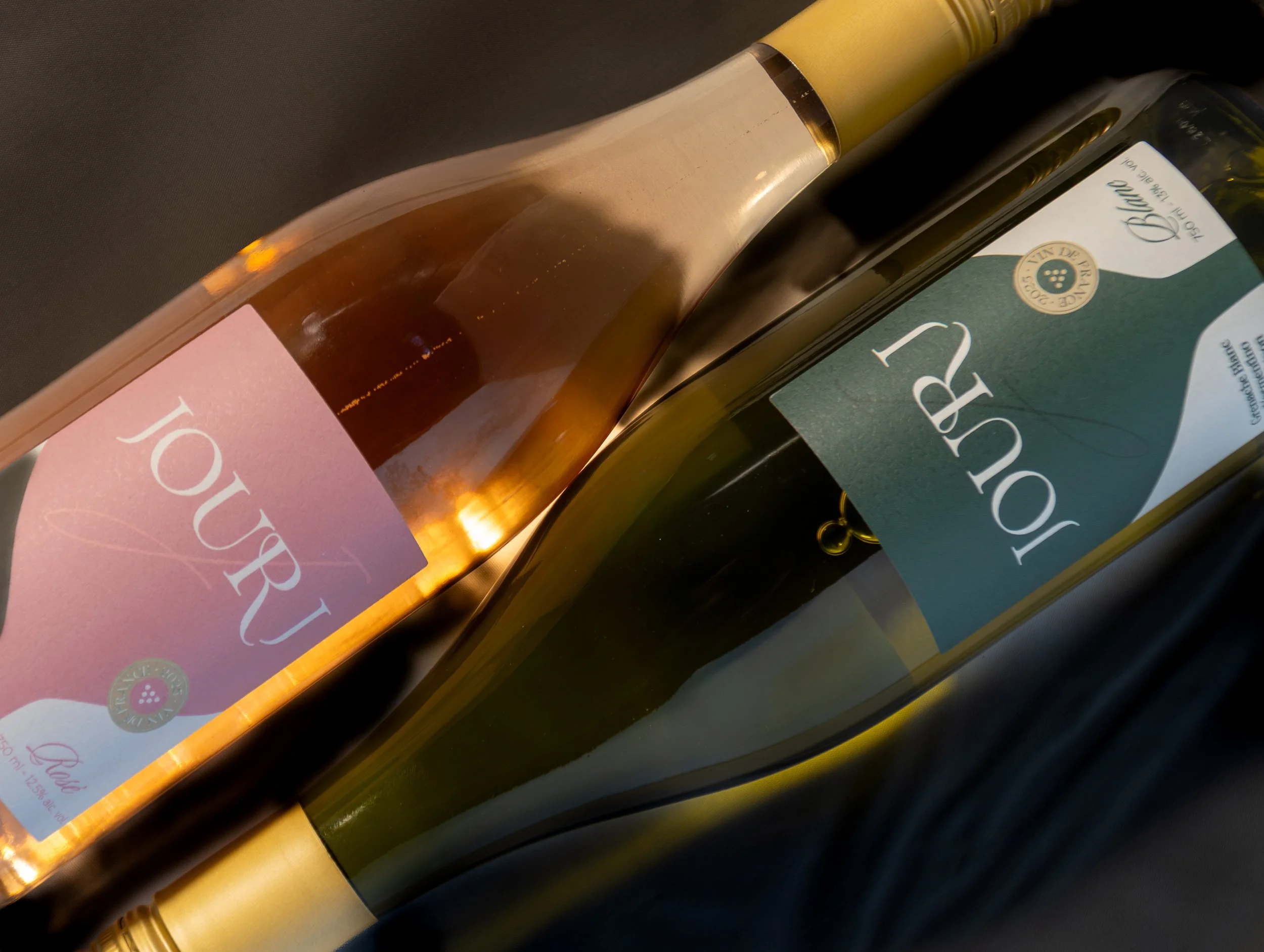 Two bottles of wine lying on a dark surface, one with a pink label reading 'TOUR' and the other with a green label reading 'TOUR'. The bottles have gold caps.