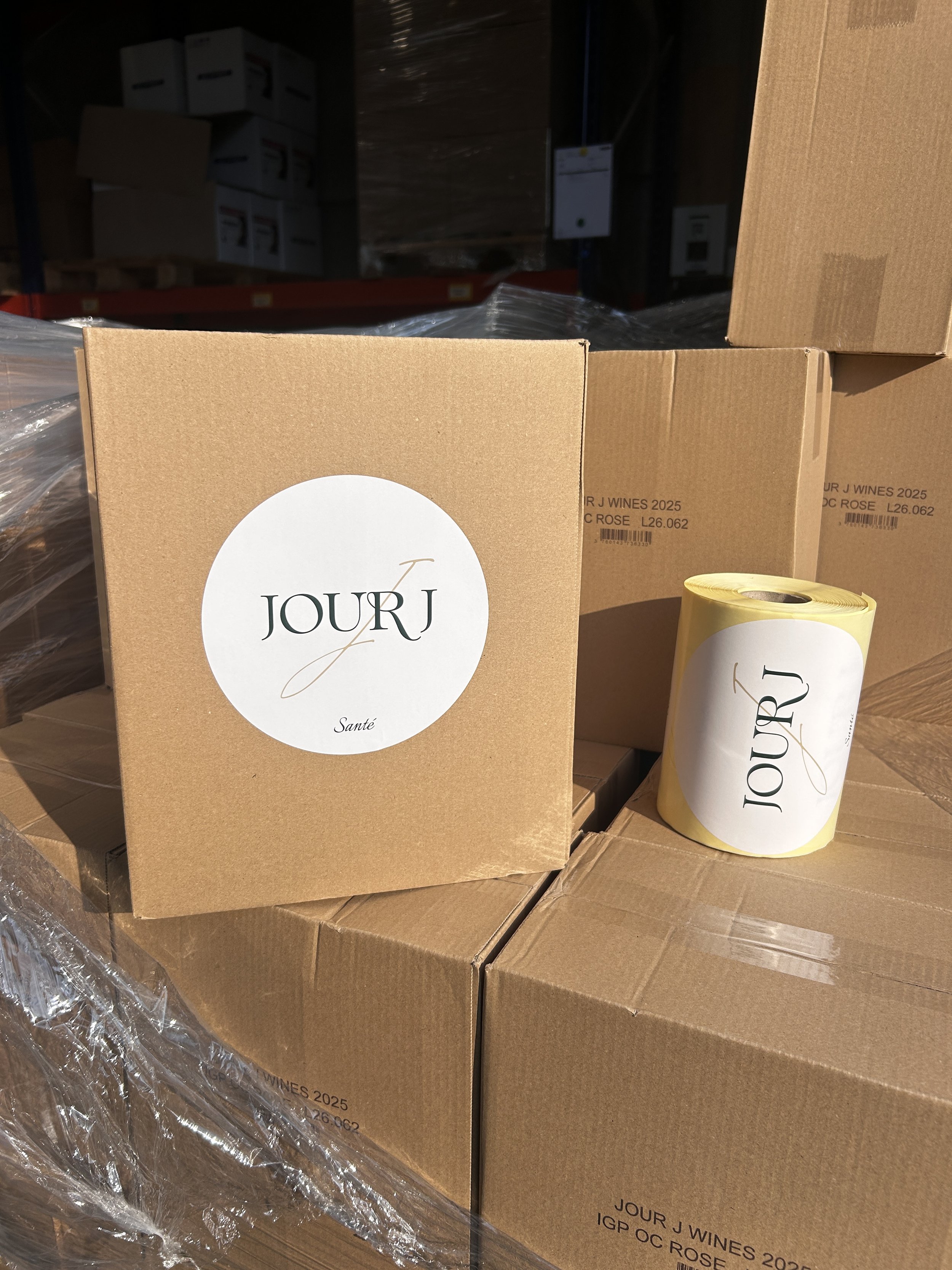 Boxes and a roll of labels with the brand name 'JOUR J' printed on them, stacked in a warehouse setting.