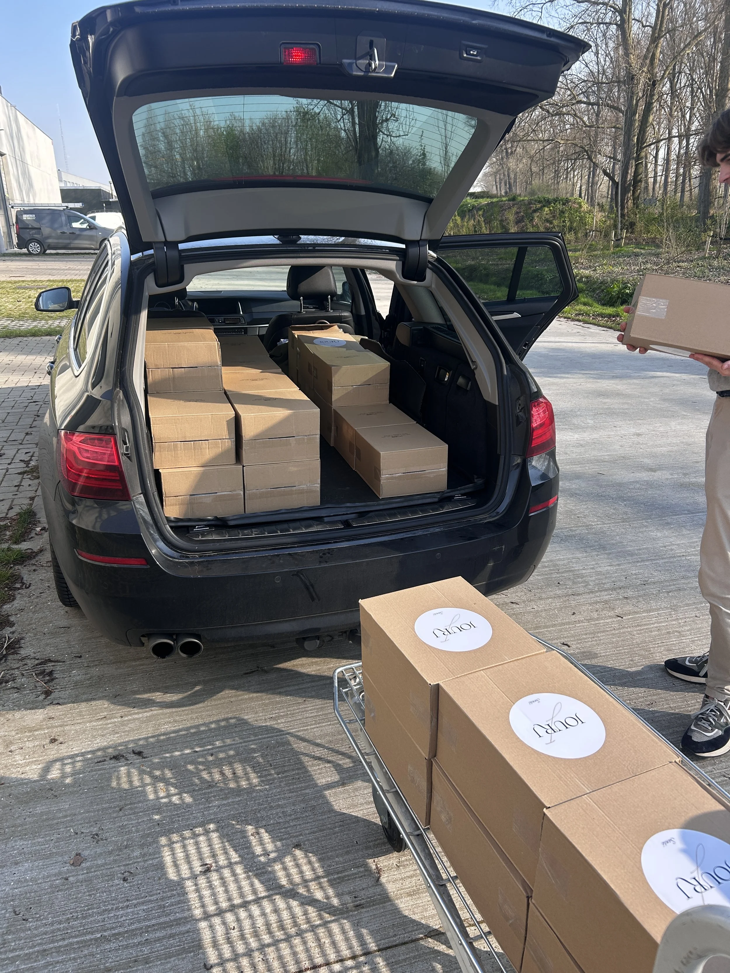 A person loading named boxes onto a car's open trunk in an outdoor parking area.