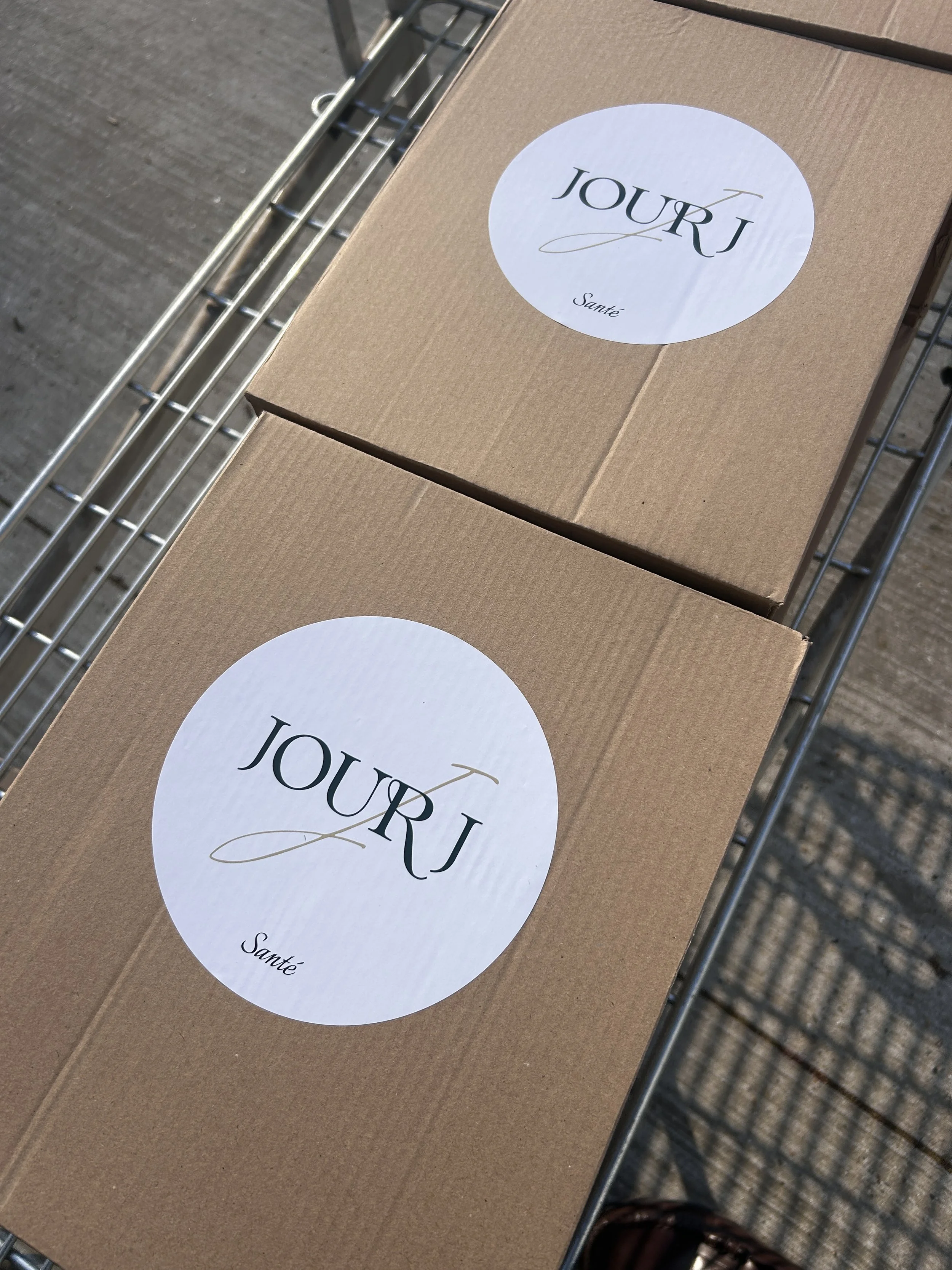 Two cardboard boxes with white circular labels reading 'JOUR' in black text, placed on a metal shopping cart.