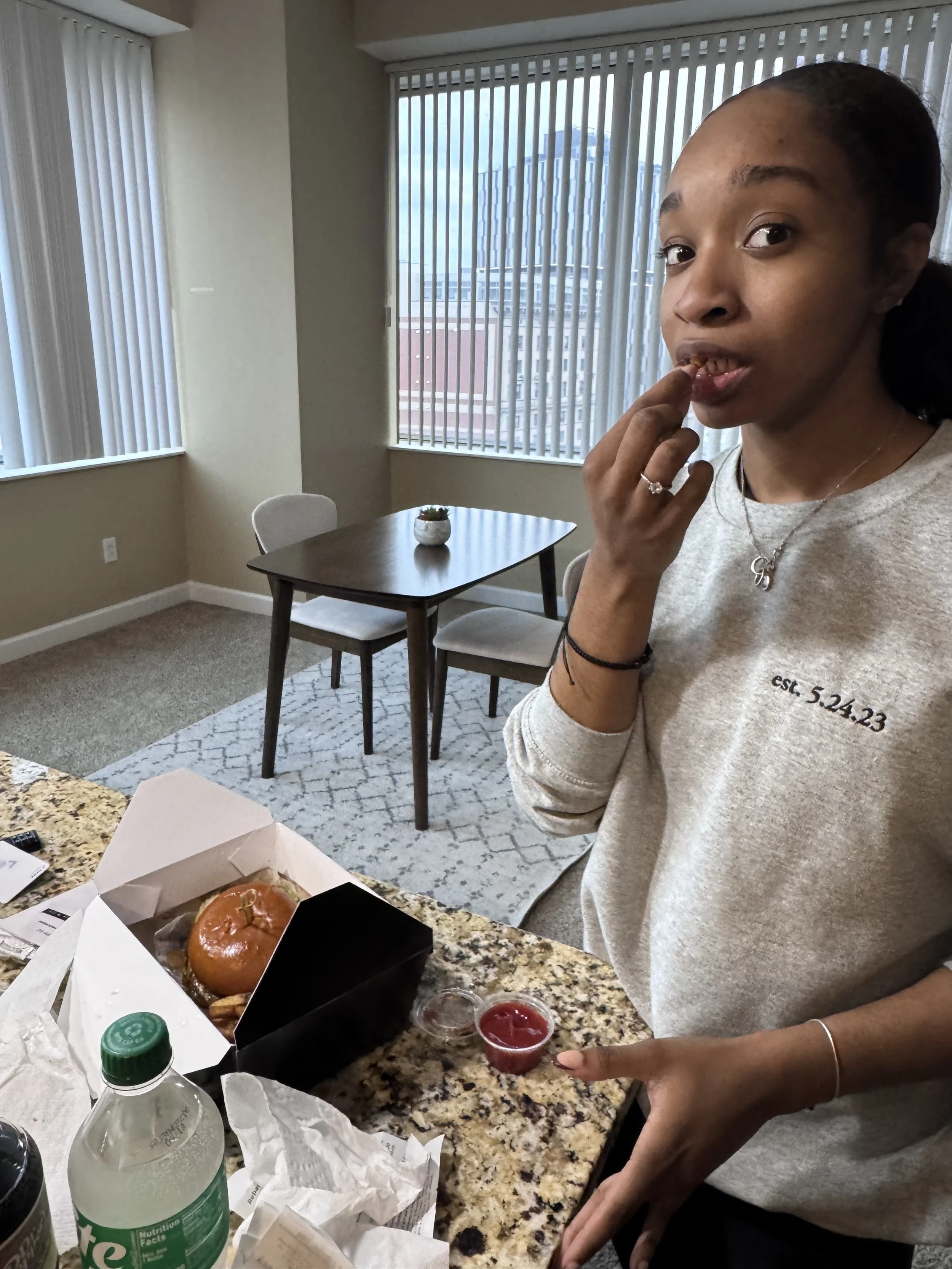 A woman standing in a kitchen with a burger in a black box on the granite countertop, a small cup of ketchup, a green bottled soda, and crumpled napkins. She has dark hair tied back, wears a light gray sweatshirt with the date 'est. 5.24.23' on it, a