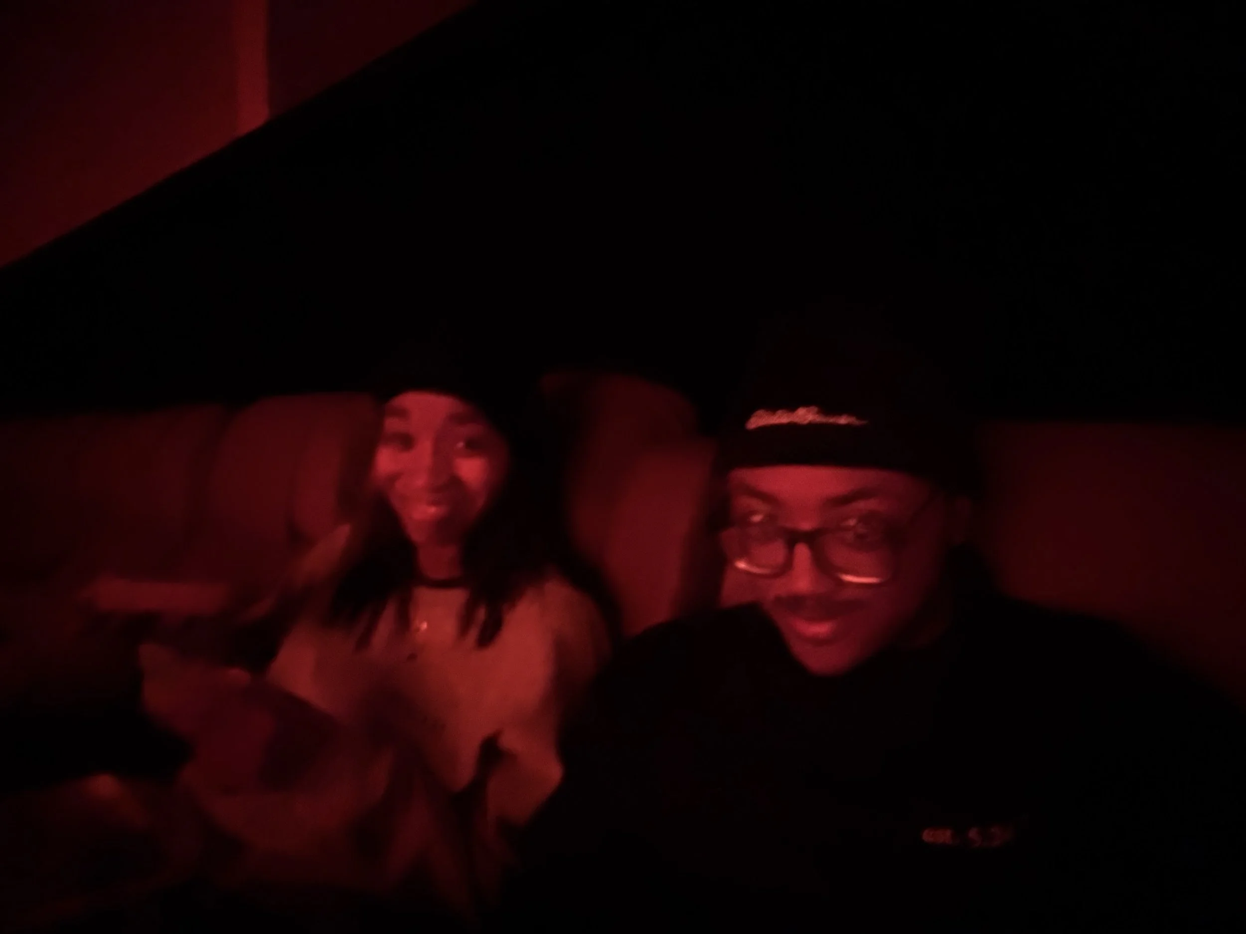 Two people smiling in a dimly lit environment with red lighting, one woman and one man, sitting on a couch