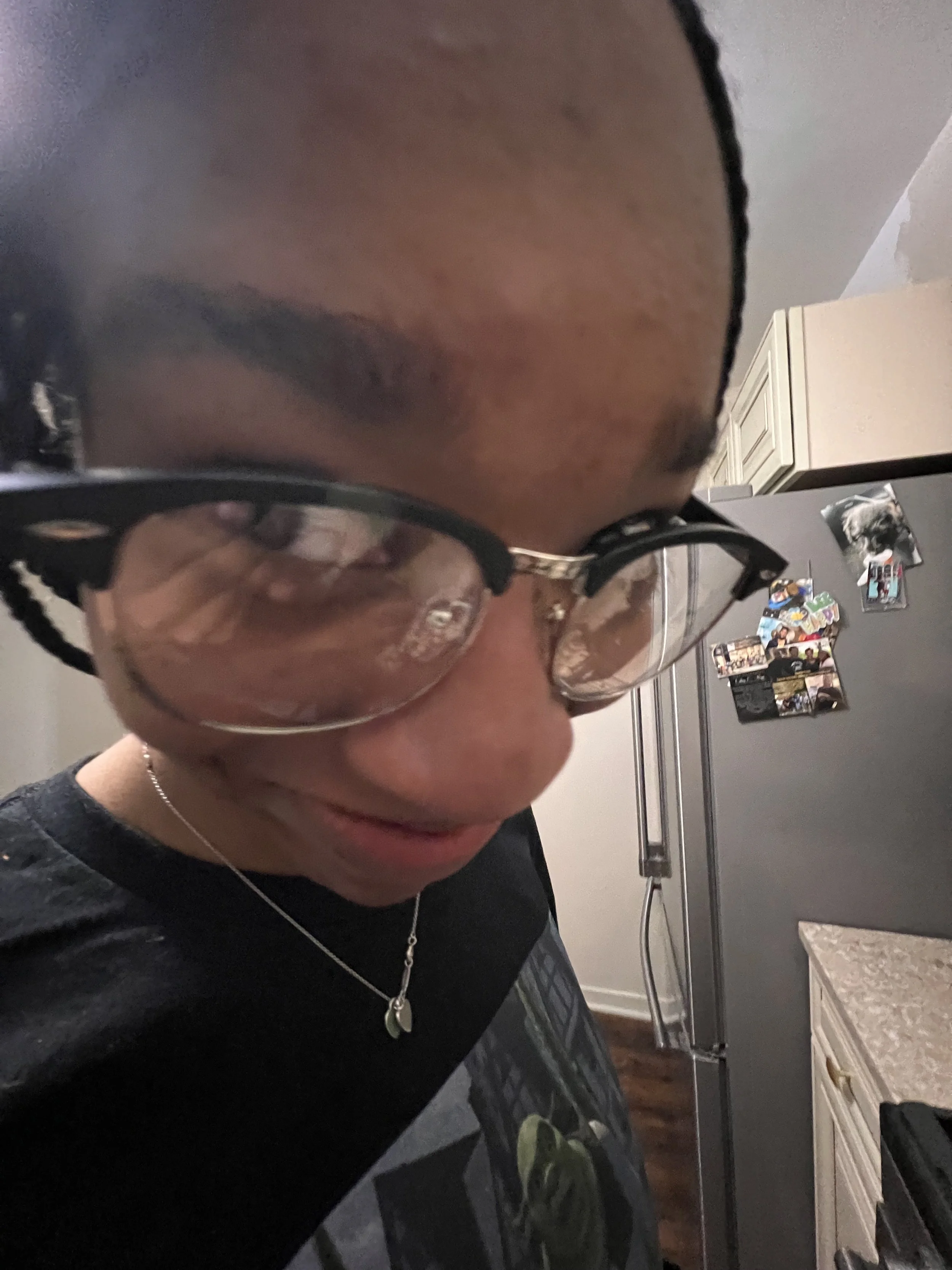A person with glasses and braided hair looking down, wearing a black t-shirt with a graphic design, a necklace, and standing in a kitchen with a refrigerator and photos on it.