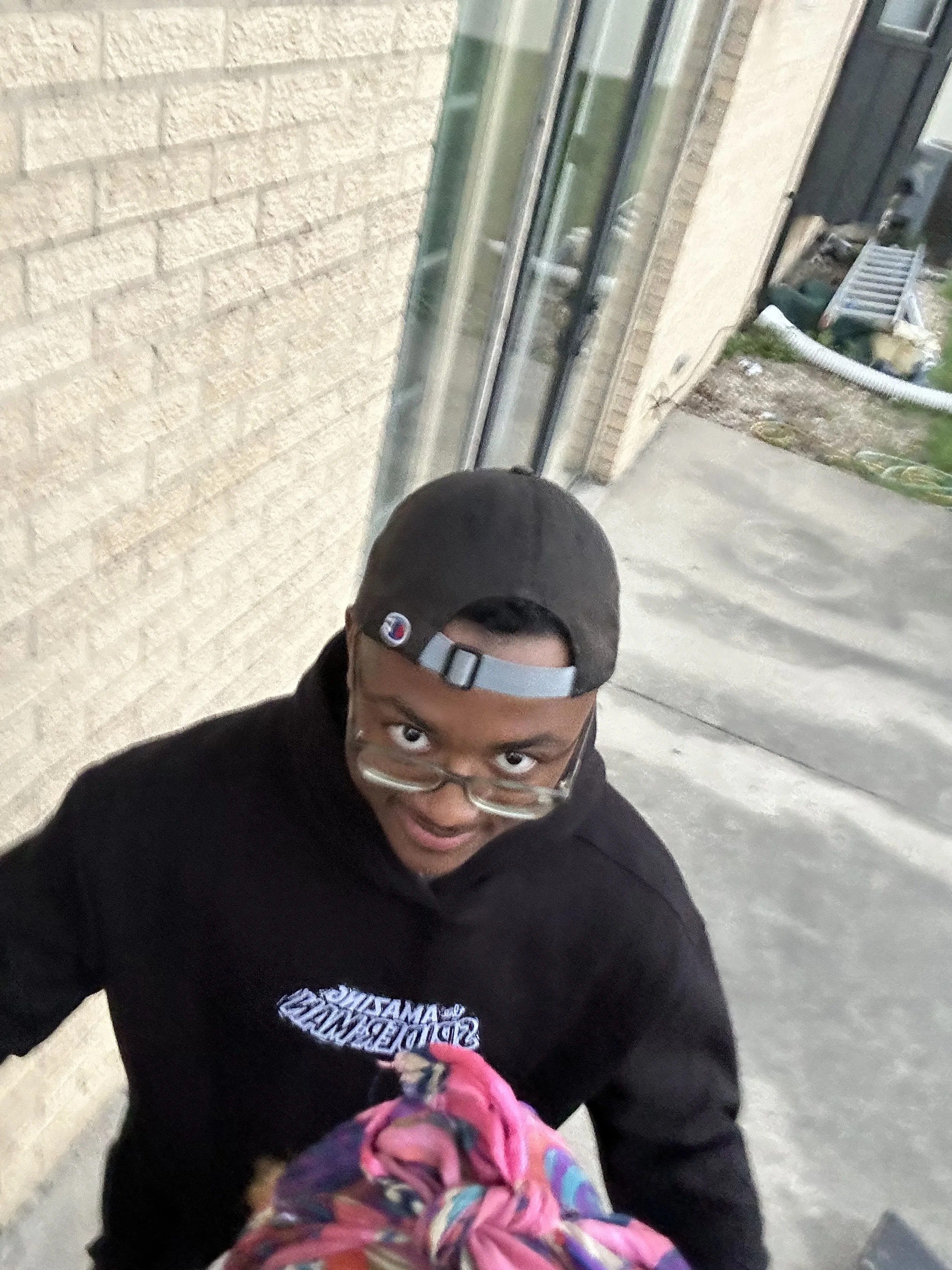 A young person with glasses, a backwards baseball cap, and a black hoodie is looking up at the camera, which is held by a person in a colorful scarf.