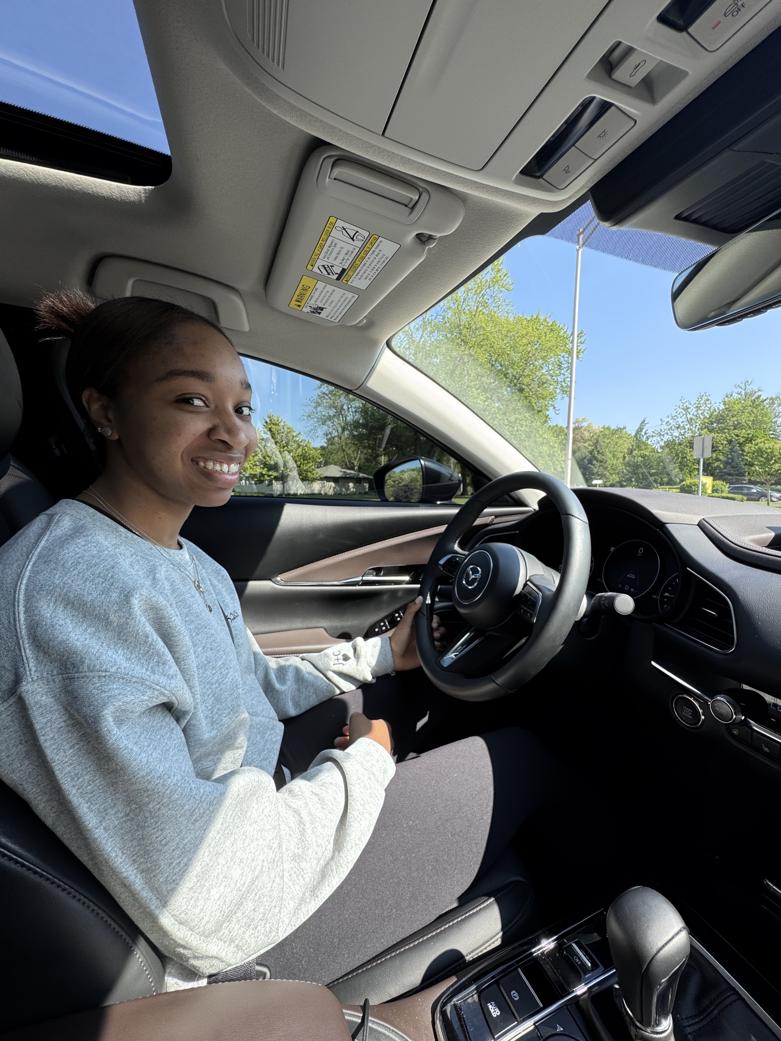 A young girl sitting in the driver's seat of a car, smiling, with her left hand on the steering wheel. The car's interior is modern, with a sunroof showing a clear blue sky and greenery outside.