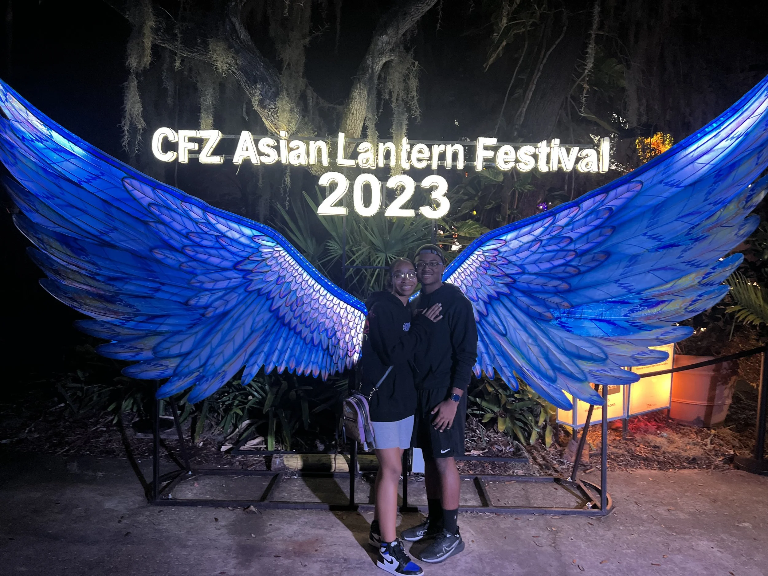 A couple standing in front of illuminated blue angel wings display at the CFZ Asian Lantern Festival 2023, nighttime setting with plants in the background.