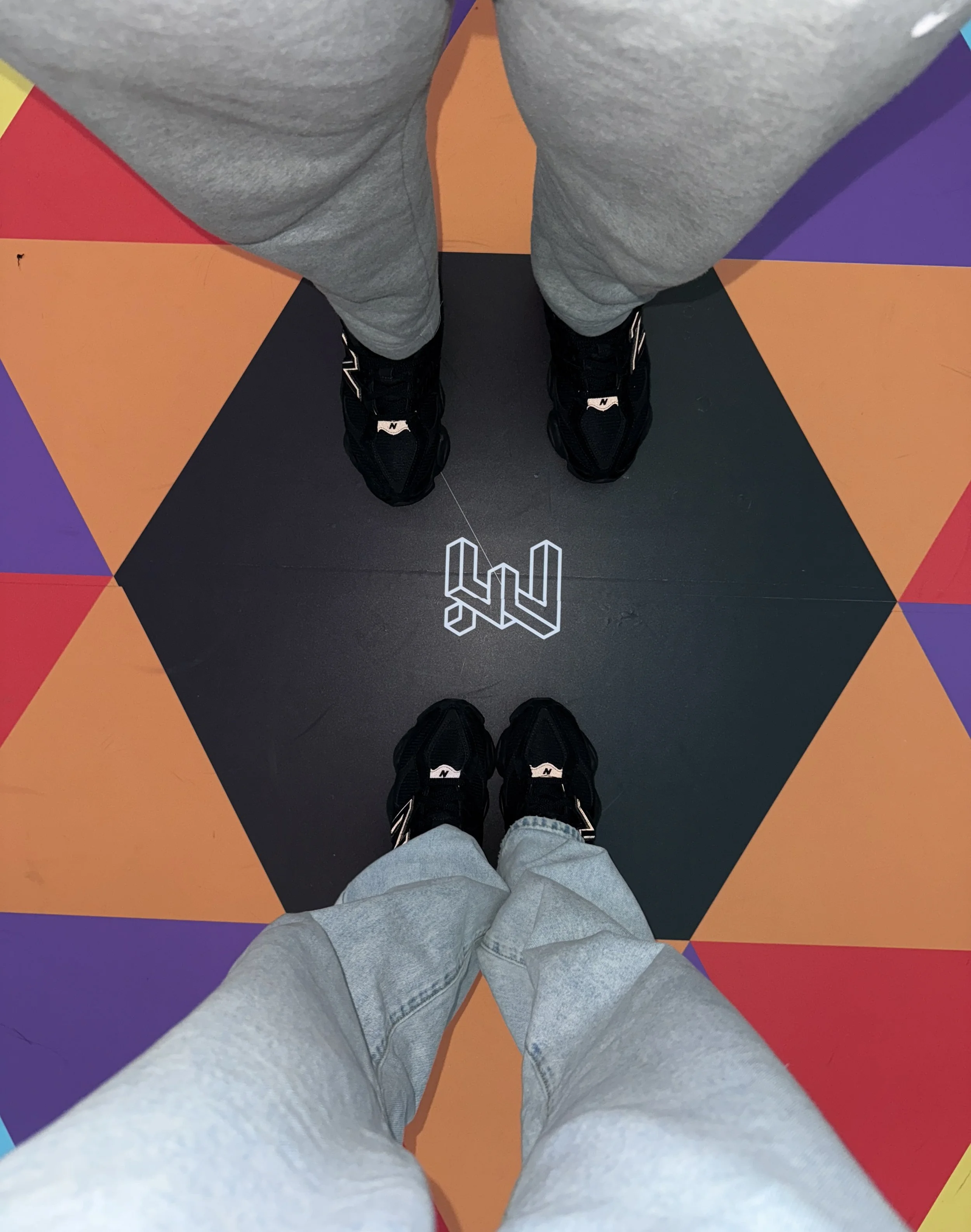 Two people standing on a colorful geometric patterned floor, both wearing black sneakers and gray sweatpants, with the logo of a fitness gym on the floor between them.