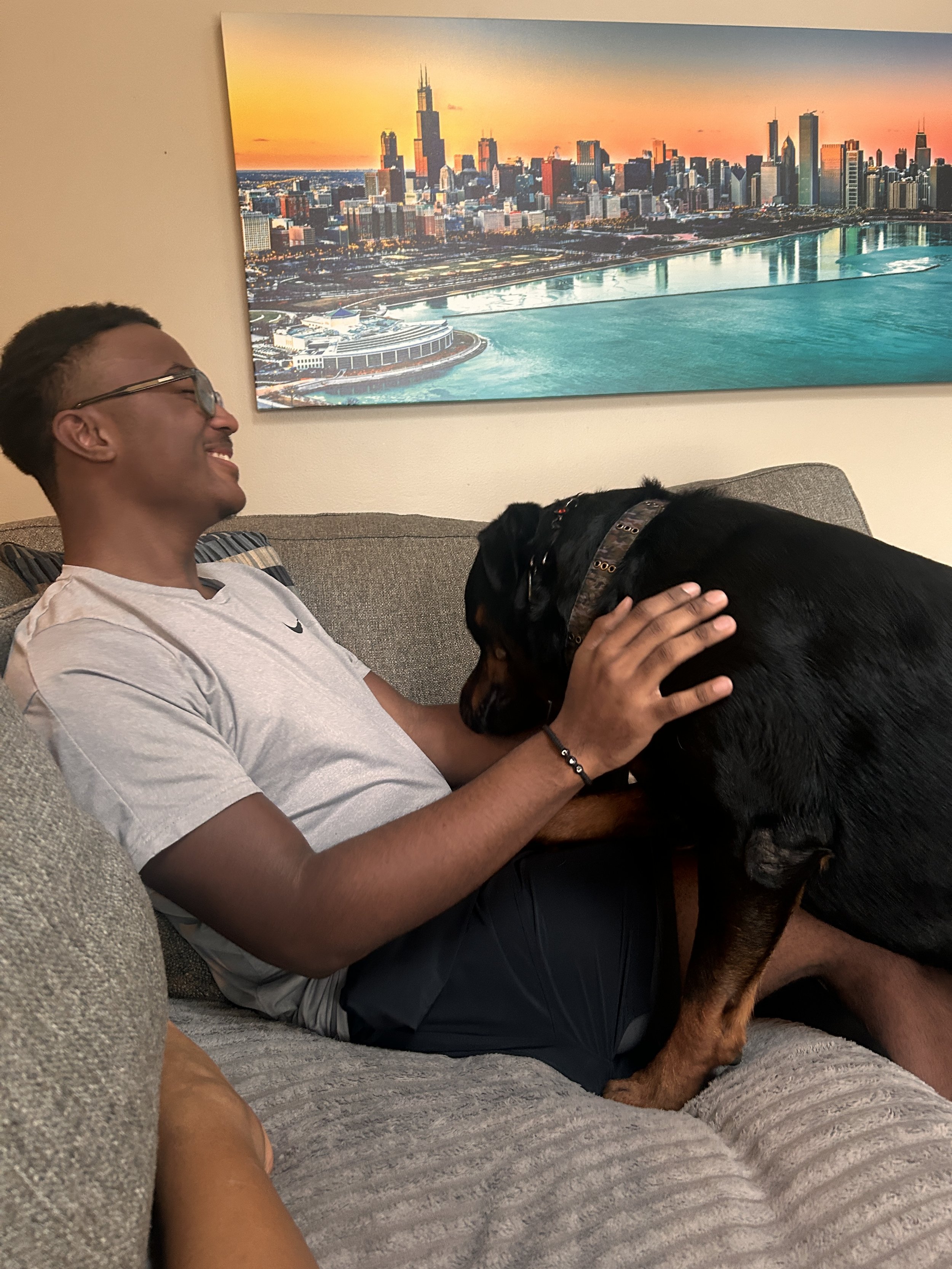 A young man with glasses and a gray t-shirt smiling and sitting on a gray couch, petting a large black and brown Rottweiler dog.