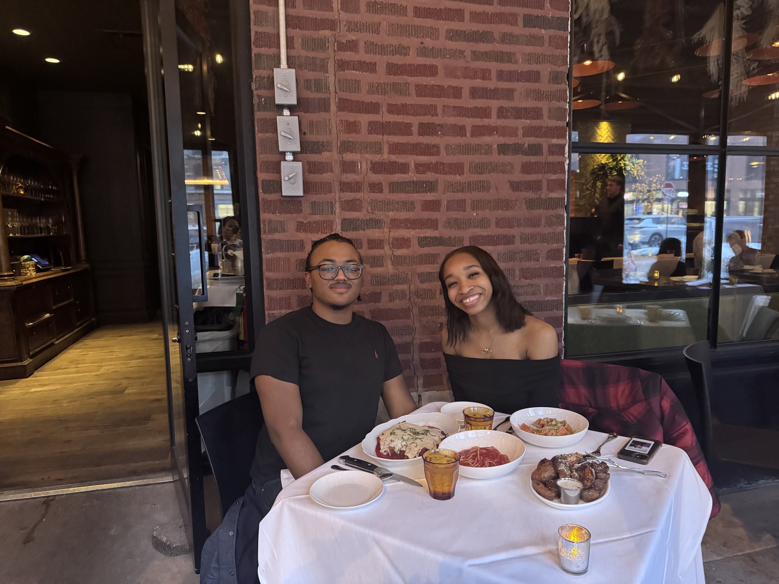 A man and a woman sit at a table in a restaurant, smiling. The table has various dishes, glasses, and utensils, with a brick wall behind them and a window showing the outside.