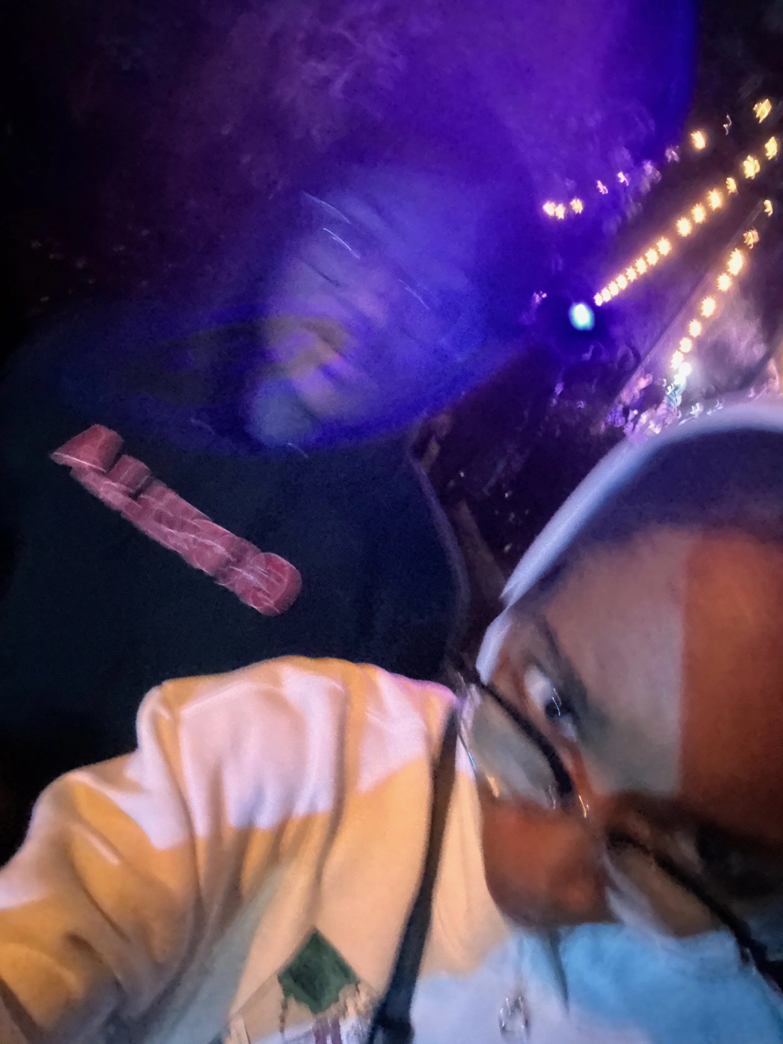 A person wearing glasses and a cap taking a selfie at an amusement park ride at night. The ride has purple and blue lights, and the person appears to be excited.