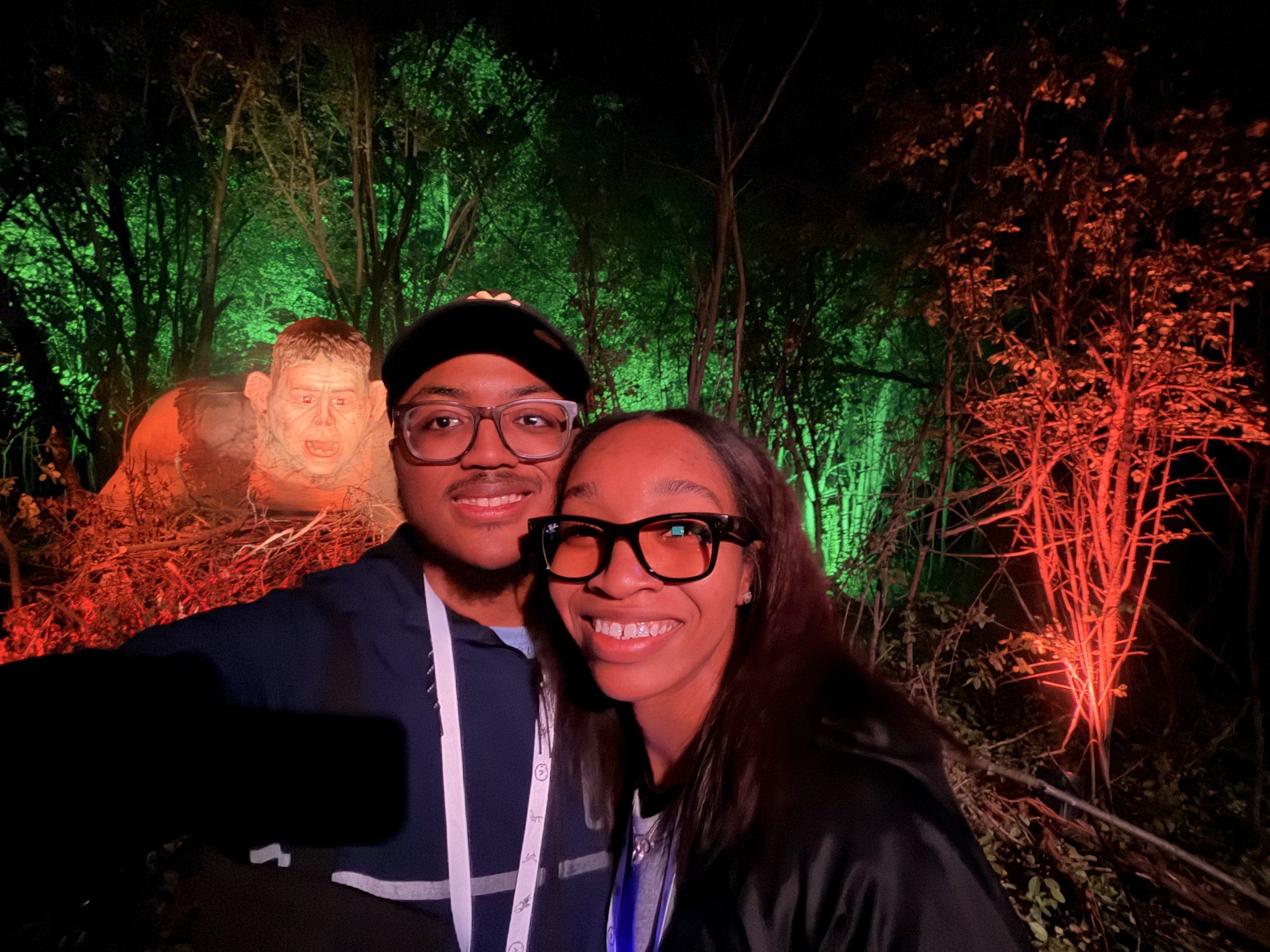 Two people, a man and a woman, smiling and taking a selfie in a dark forest with vibrant green and red lights illuminating the trees. There is an orange inflatable figure with a painted face in the background.