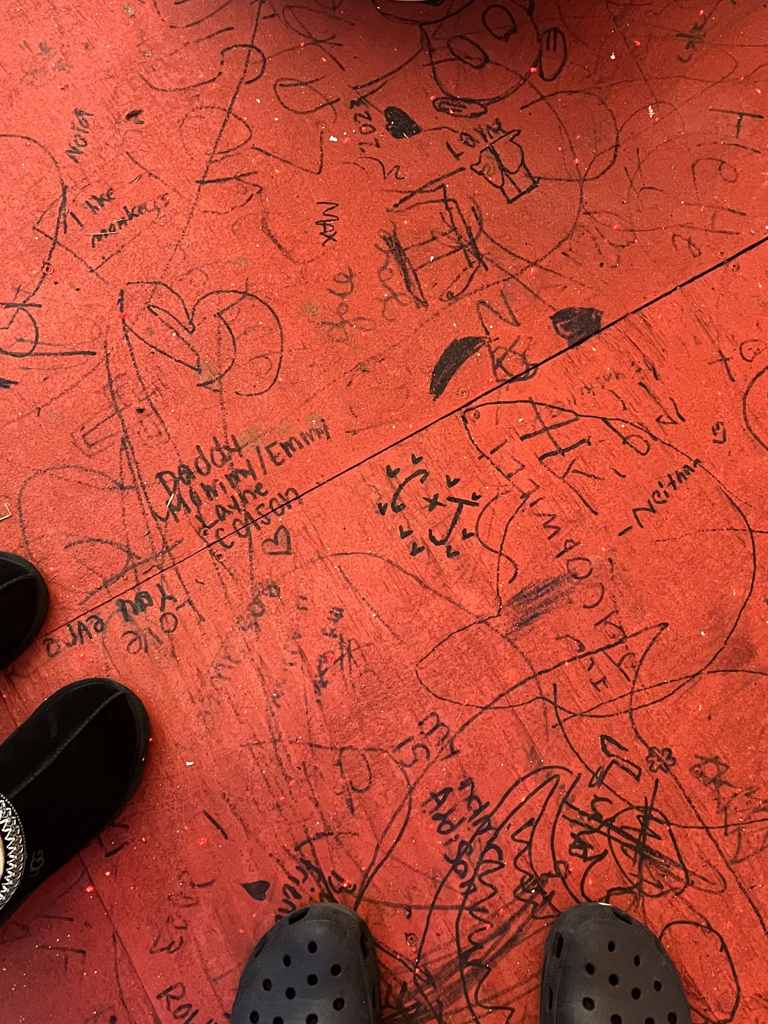 Red floor covered with black ink doodles, signatures, and messages, with the feet of two people in Crocs shoes visible at the bottom.