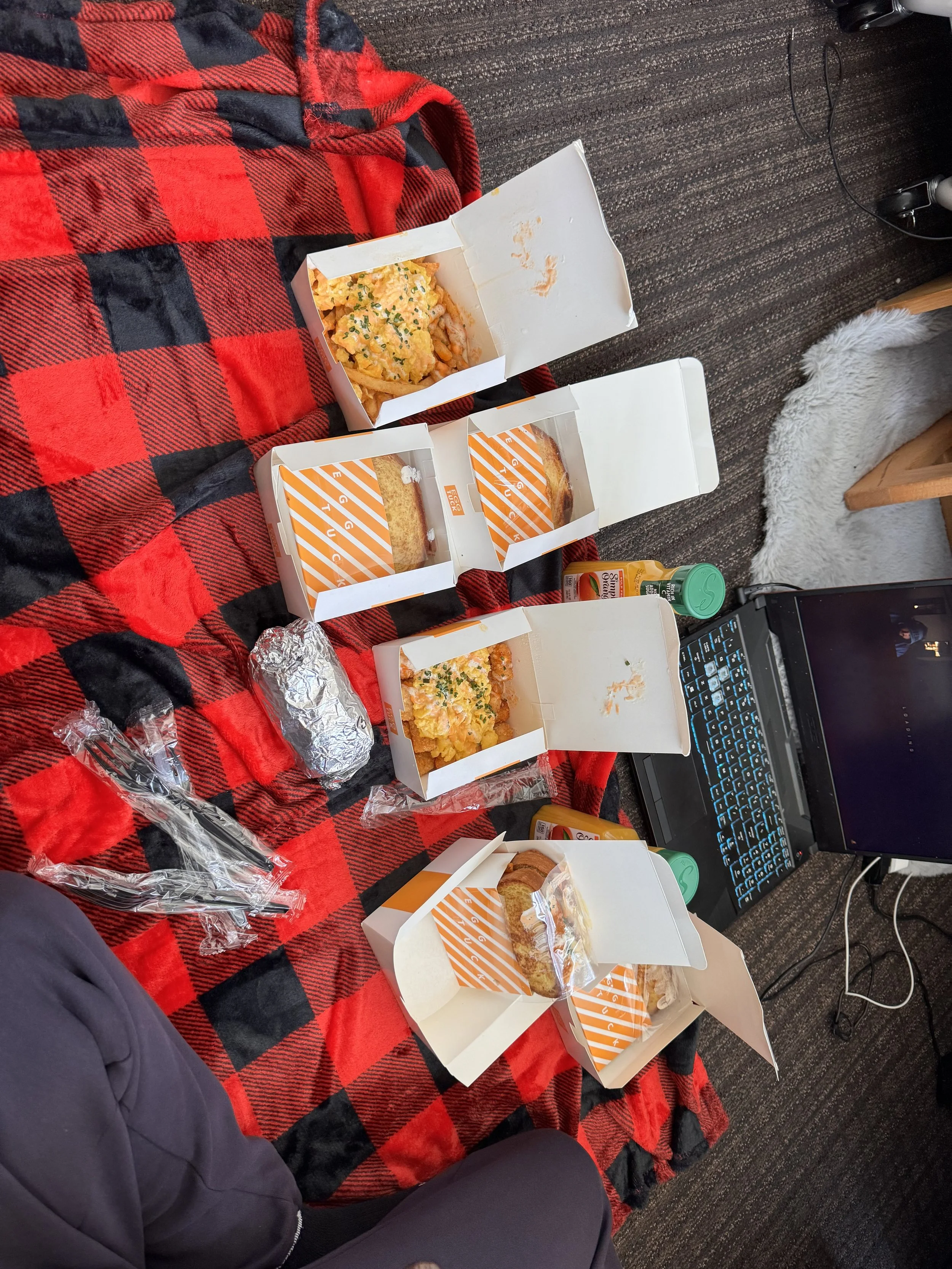 Several open fast food boxes with fried food on a red and black checkered table covering. Some wrapped items and plastic utensils are also on the table next to a laptop, condiments, and a person's leg.