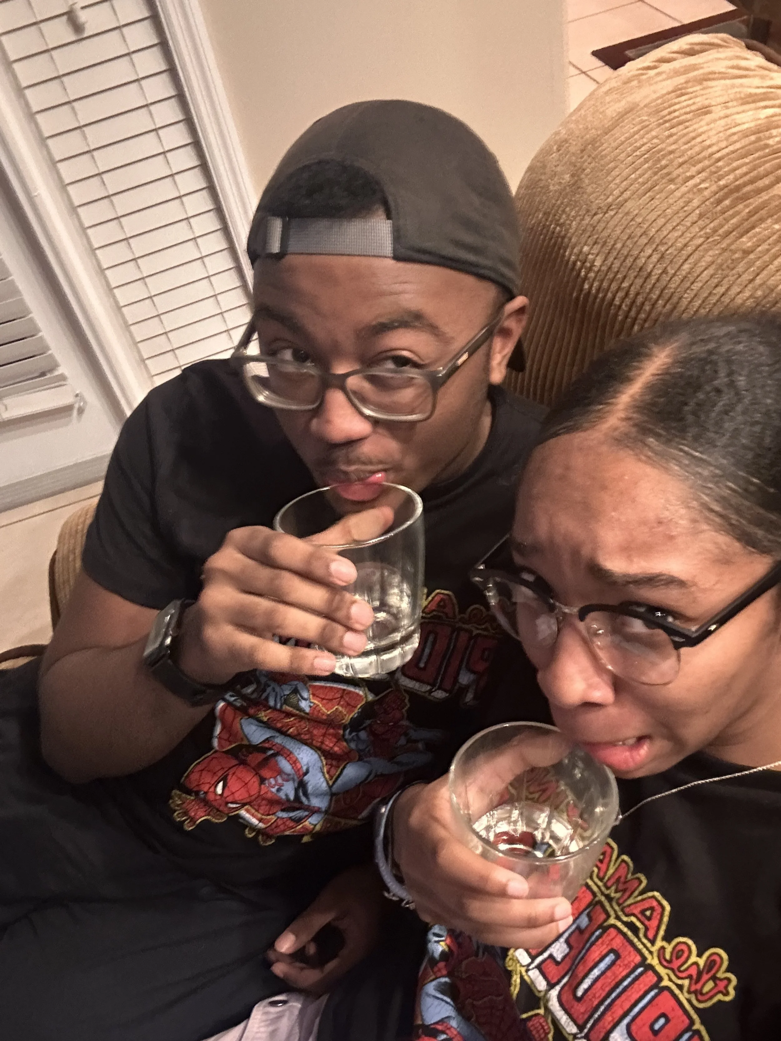 Two young adults, a man and a woman, sitting close together (possibly on a couch), each holding a glass of clear liquid. They are wearing glasses and matching Spider-Man themed shirts. The man is wearing a backwards cap.