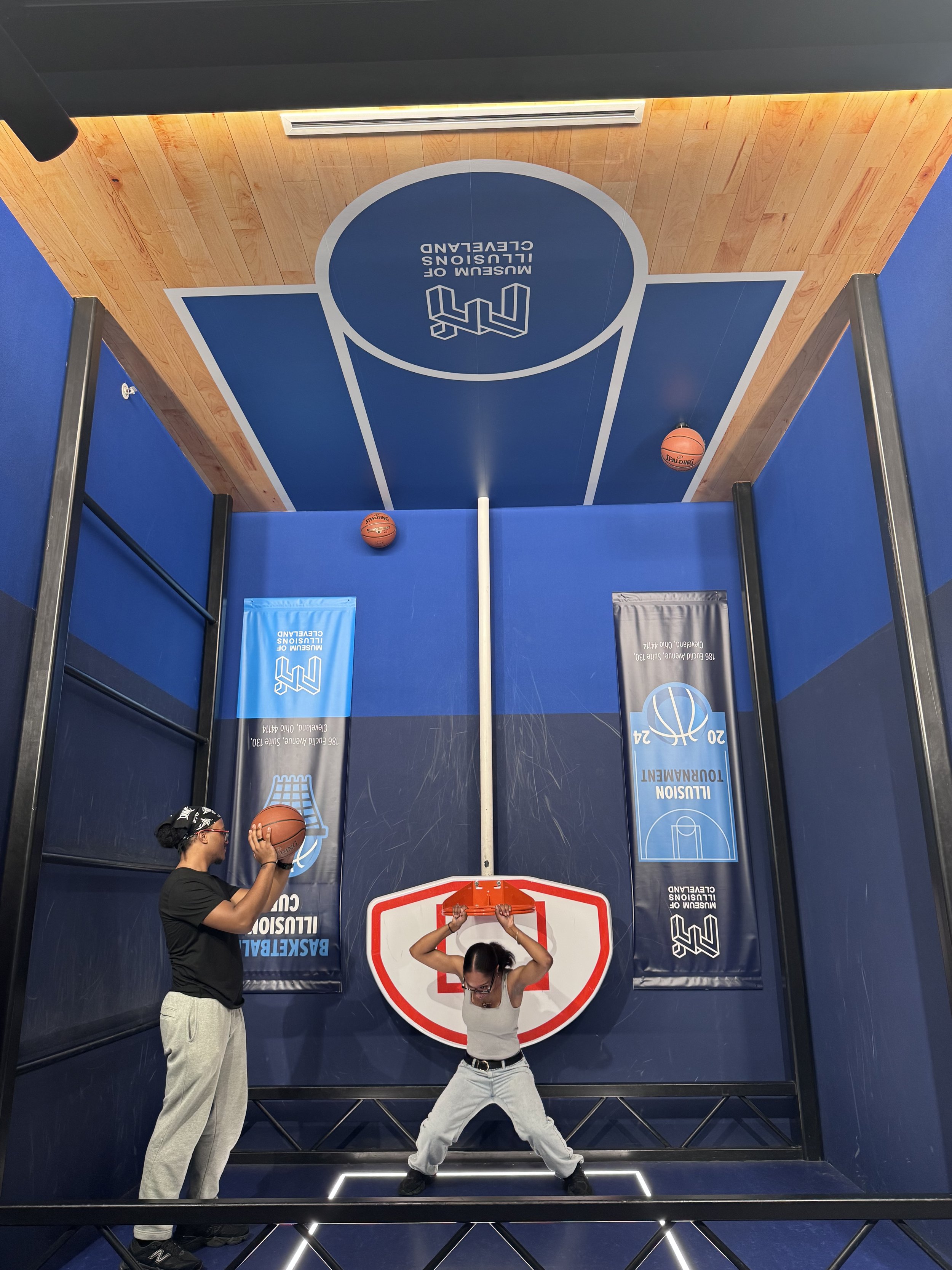 Two women playing basketball on an indoor court with blue walls and a wooden ceiling, one preparing to shoot and the other holding a basketball.