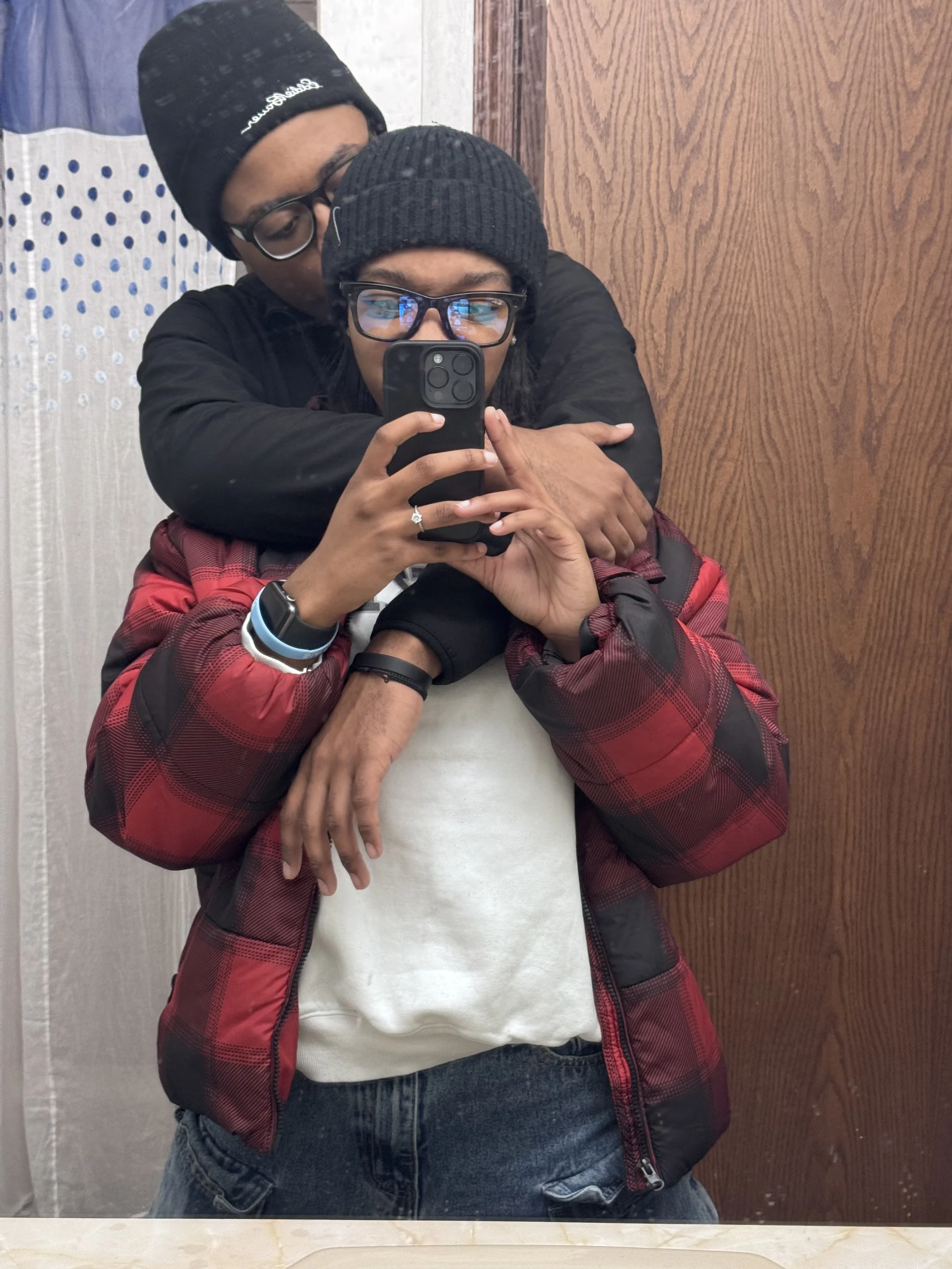 Two young people taking a mirror selfie together in an indoor setting. The person in front is holding a smartphone and wearing glasses, a black beanie, a red plaid jacket, and a white shirt, with a smartwatch on their left wrist. The person behind we