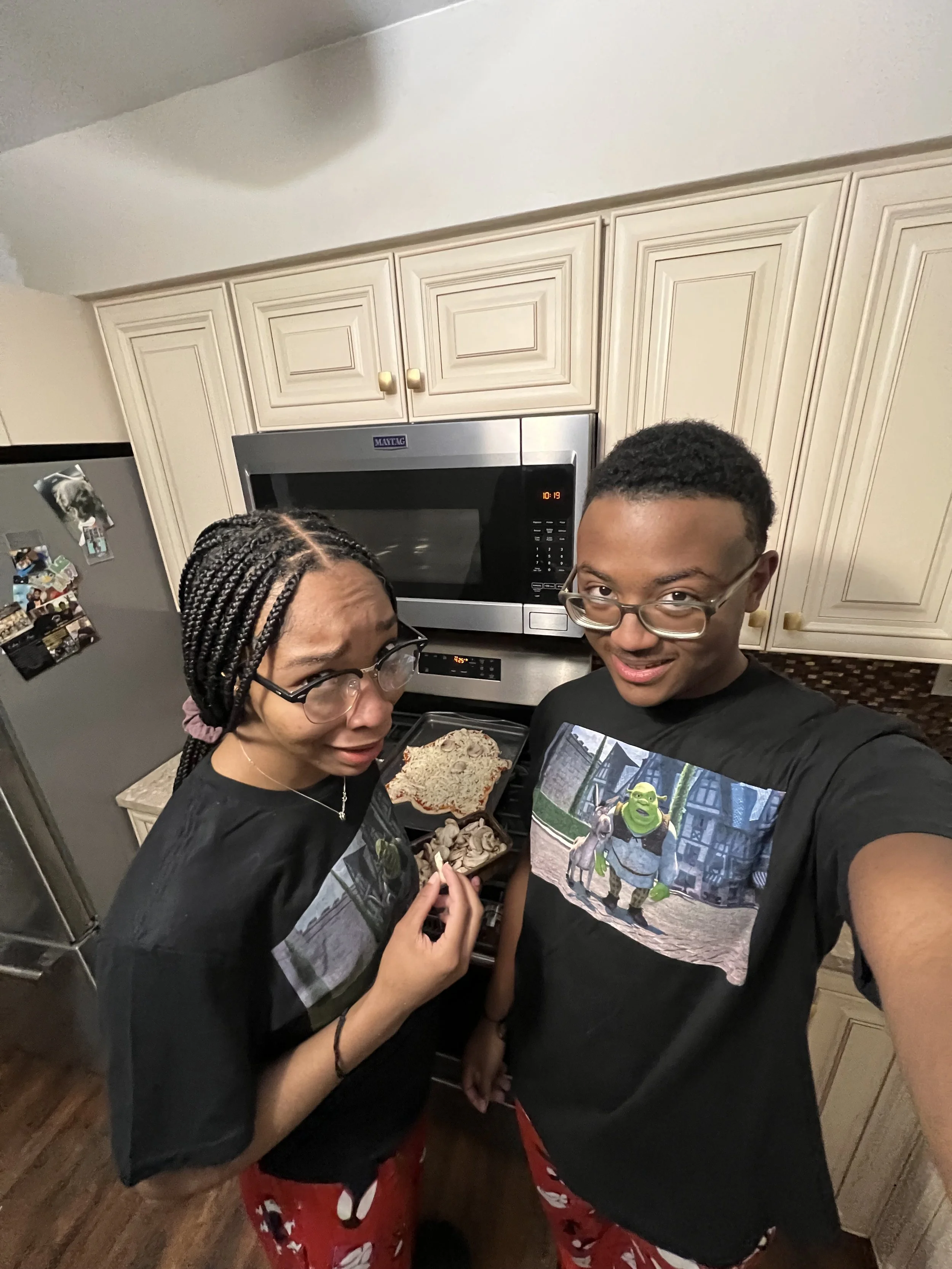 A young man and woman in a kitchen, both wearing glasses and black T-shirts with animated characters, standing close together and smiling at the camera. There is a microwave, a fridge with photos, and a tray with a pizza and some snacks on the stove behind them.
