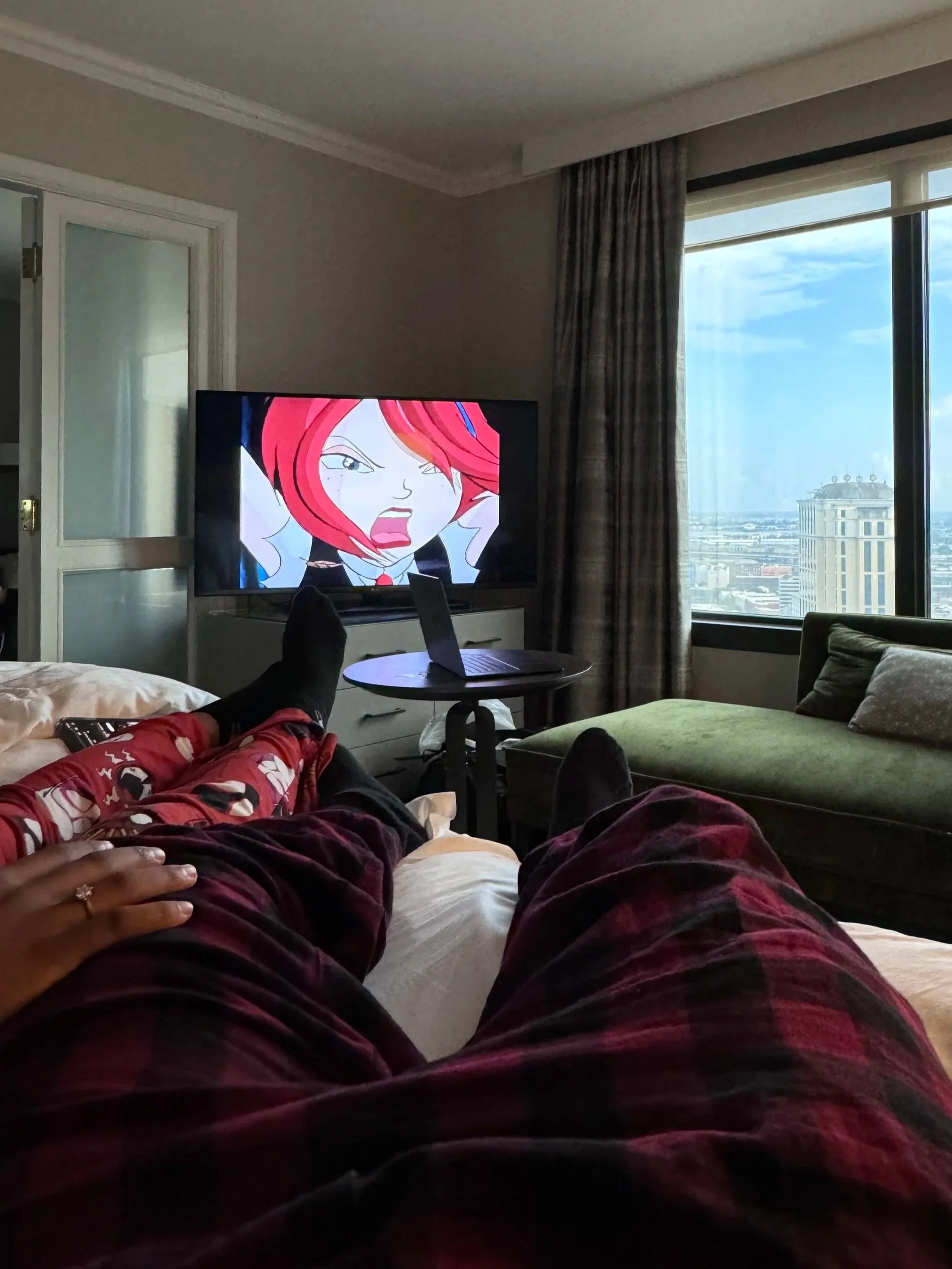 A person lying on a bed with legs stretched out, watching an animated show on a television in a hotel room with large windows view of city buildings in the background.