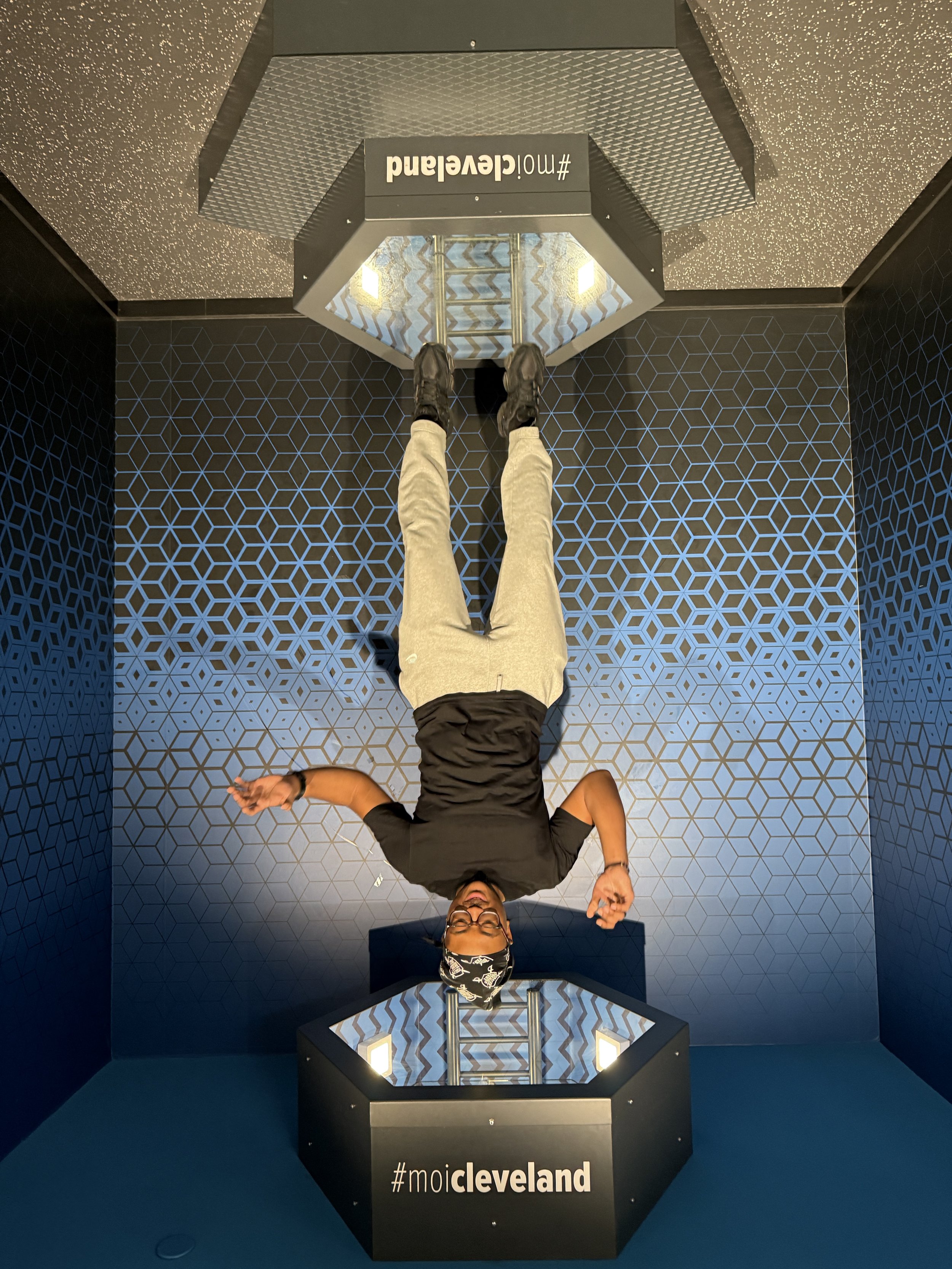 A person lying on the floor in a room with black walls and a geometric blue pattern. The person is wearing a black shirt, beige pants, black shoes, glasses, and a patterned bandana. They are smiling and making a hand gesture. There are two hexagonal 