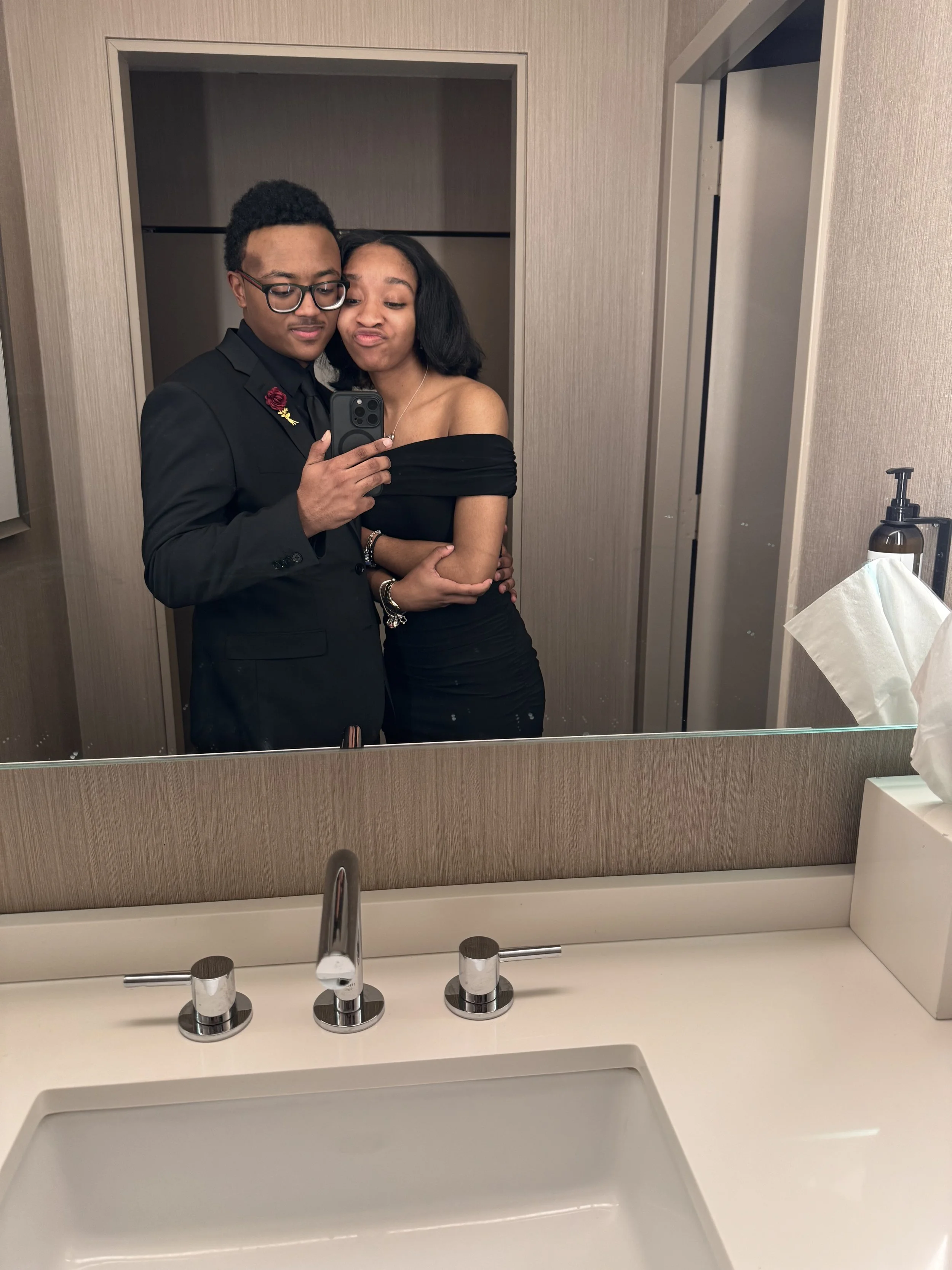 A young man and woman taking a selfie in front of a bathroom mirror. The man is dressed in a black suit with a red rose lapel pin and glasses. The woman, in a black off-shoulder dress, has short black hair and is making a pout face while hugging the man.