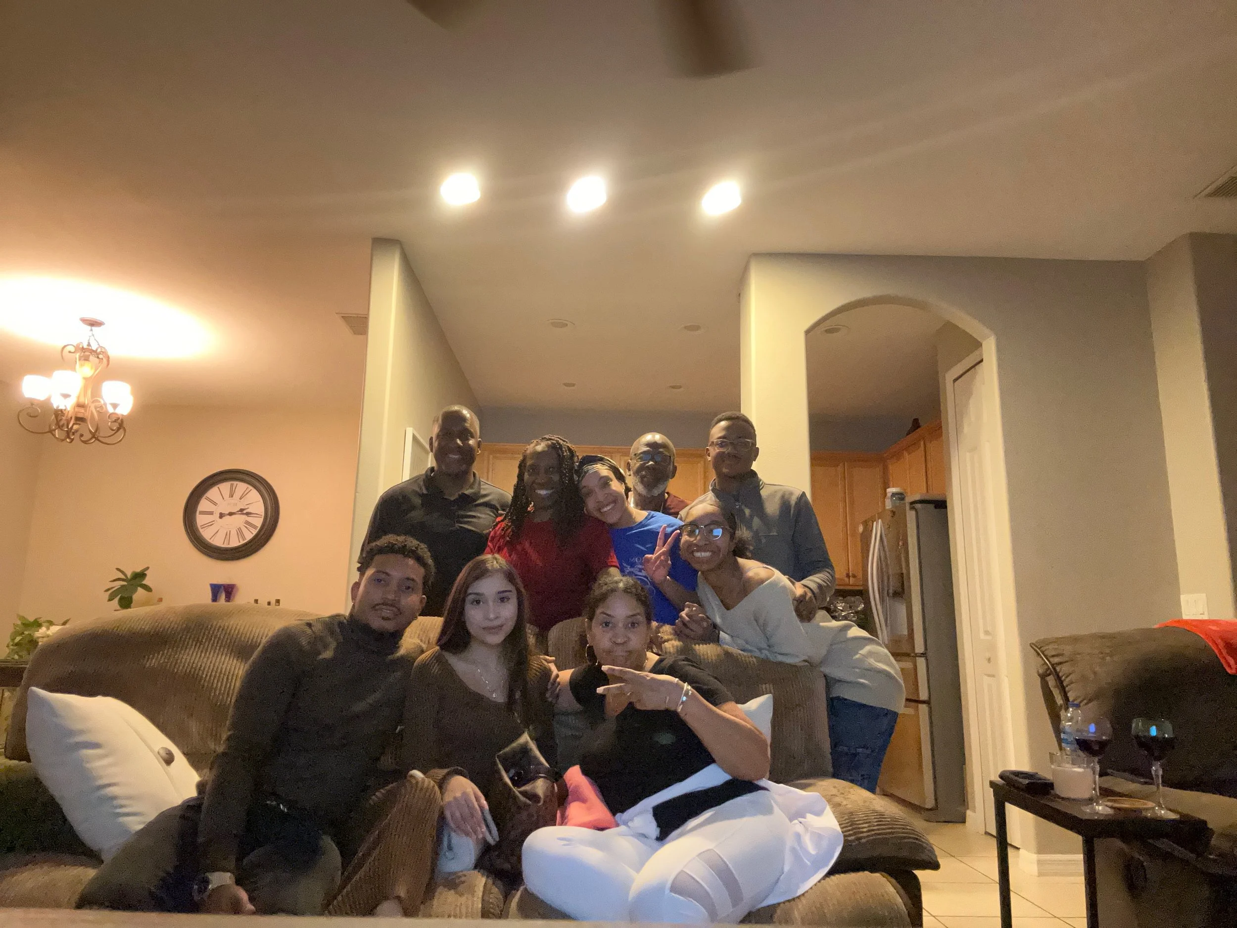 Group of ten people gathered in a living room, smiling and posing for a photo, with wooden kitchen cabinets visible in the background.