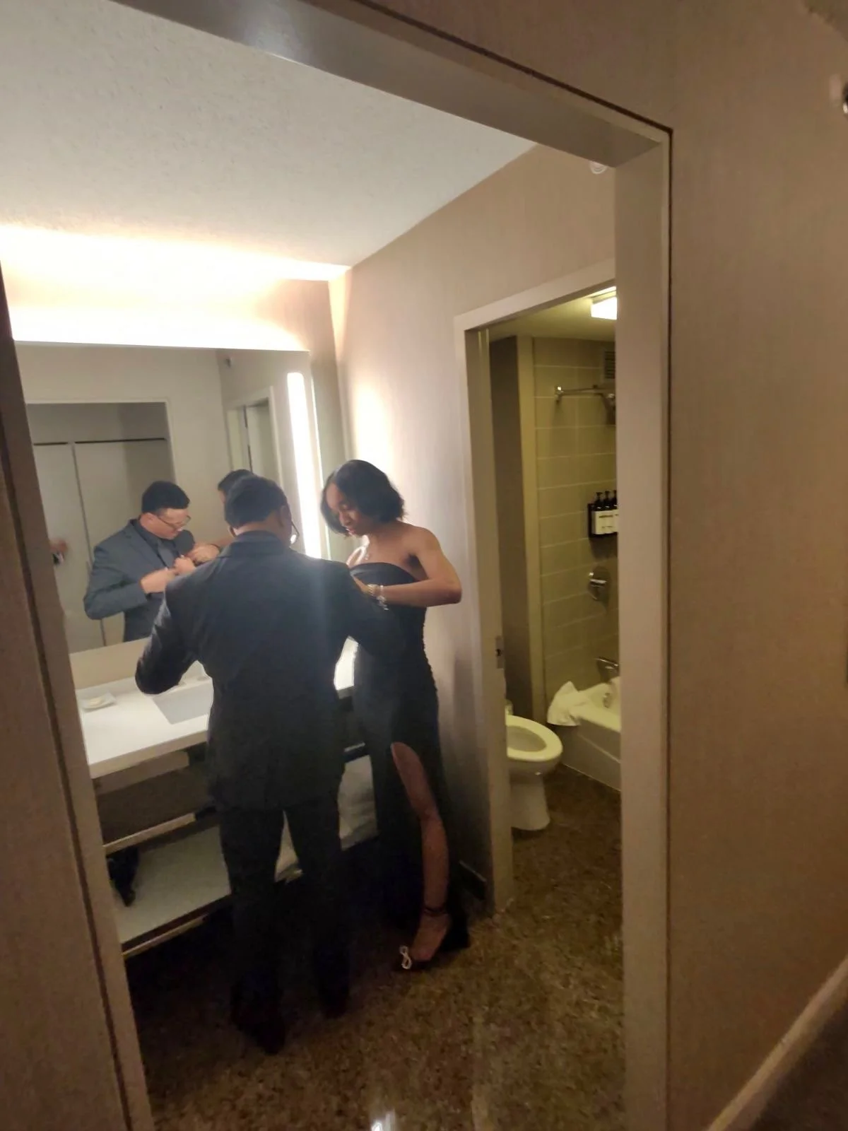 A group of three people in formal attire, two men and a woman, standing in front of a mirror in a hotel room bathroom, with a toilet visible behind them.