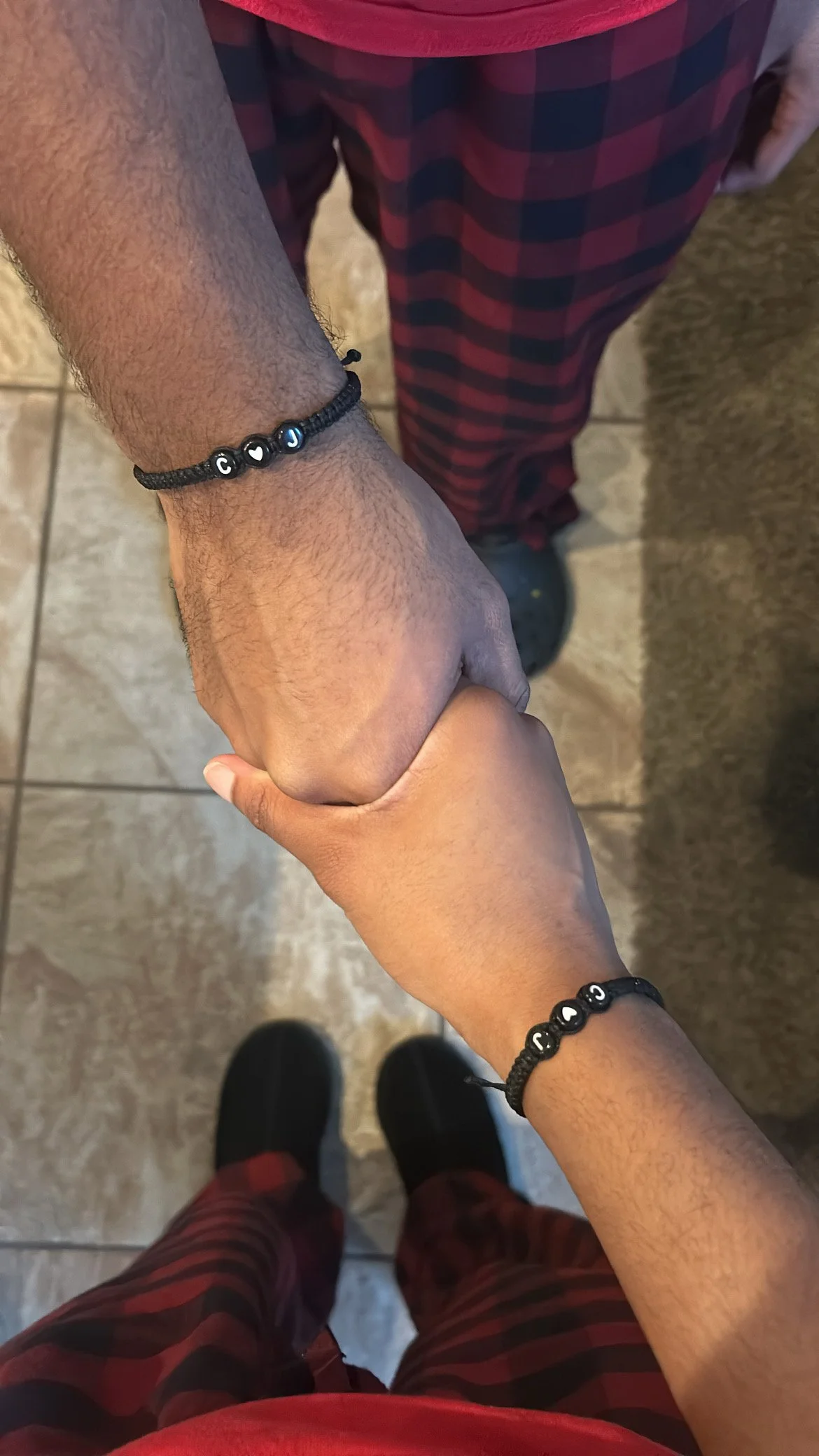 Two people holding hands, wearing matching black wristbands with letter beads. One person wears a red shirt and checkered pants, the other wears a darker shirt.