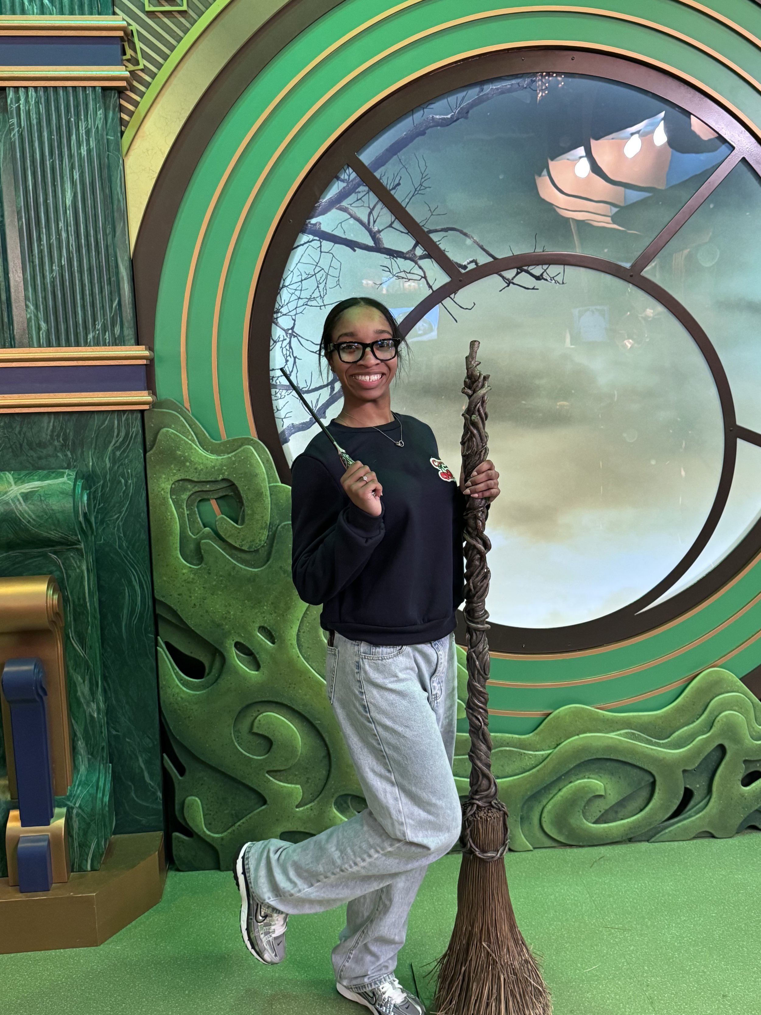 Young woman wearing glasses, black sweatshirt, and gray pants, smiling and holding a pretend magic wand and a broomstick in front of a green, fantasy-themed backdrop with a large clock window and leaf designs.