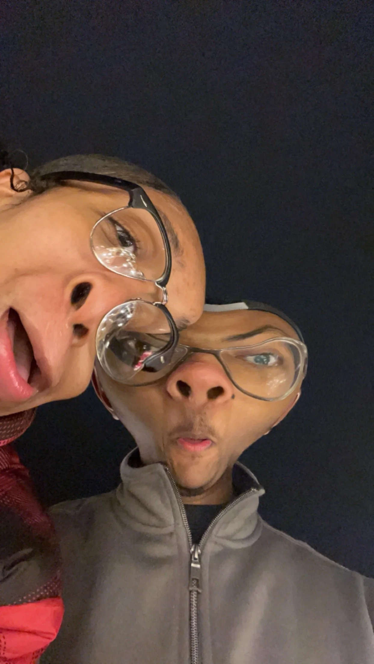 Two people making funny faces, wearing glasses, with a dark background.