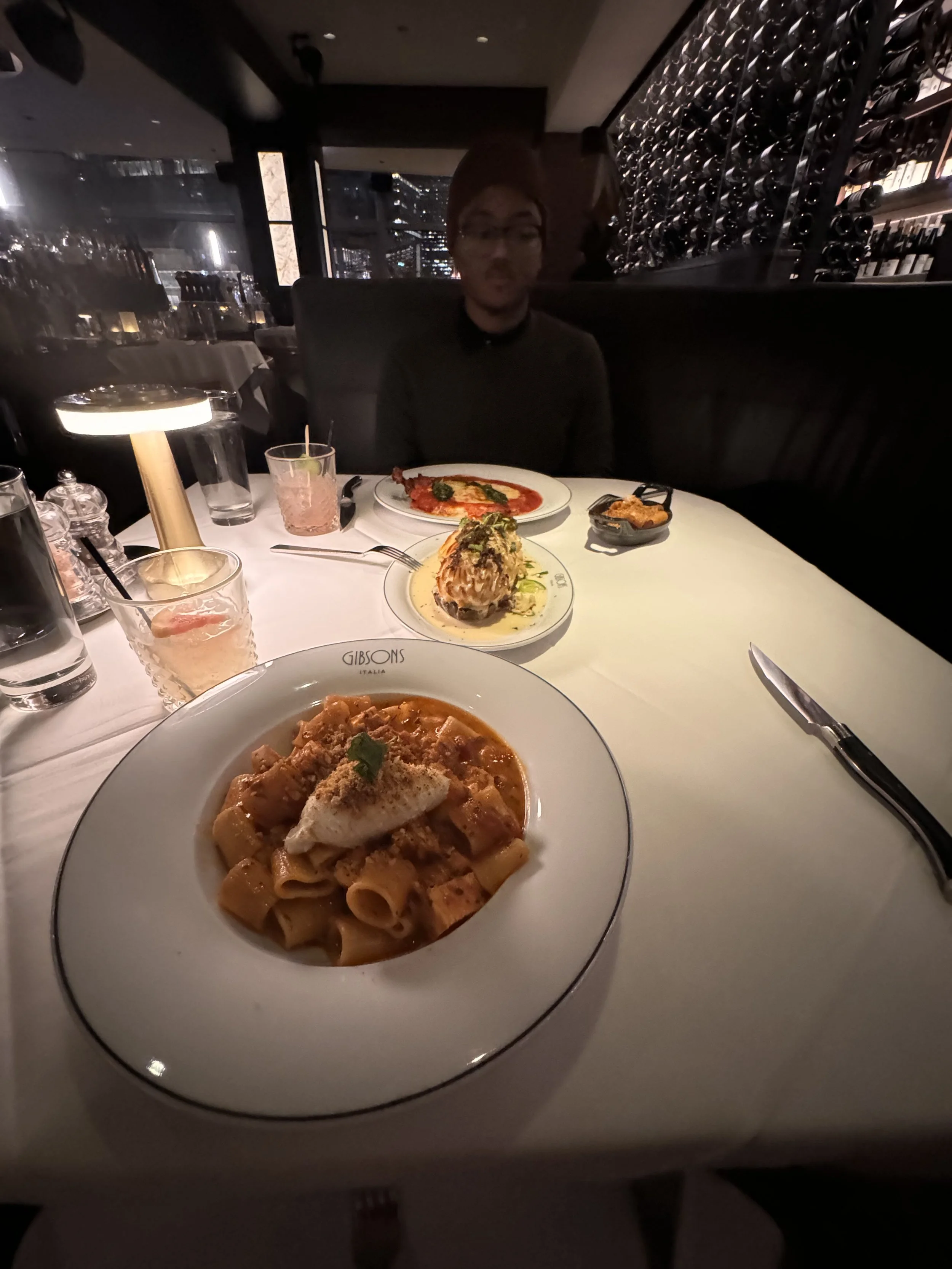 A person in a dark restaurant sitting behind a white table with three plates of food, including pasta with tomato sauce and a dollop of cream, a stuffed mushroom, and a dish with tomato slices. There are also glasses of water and pink cocktails on th