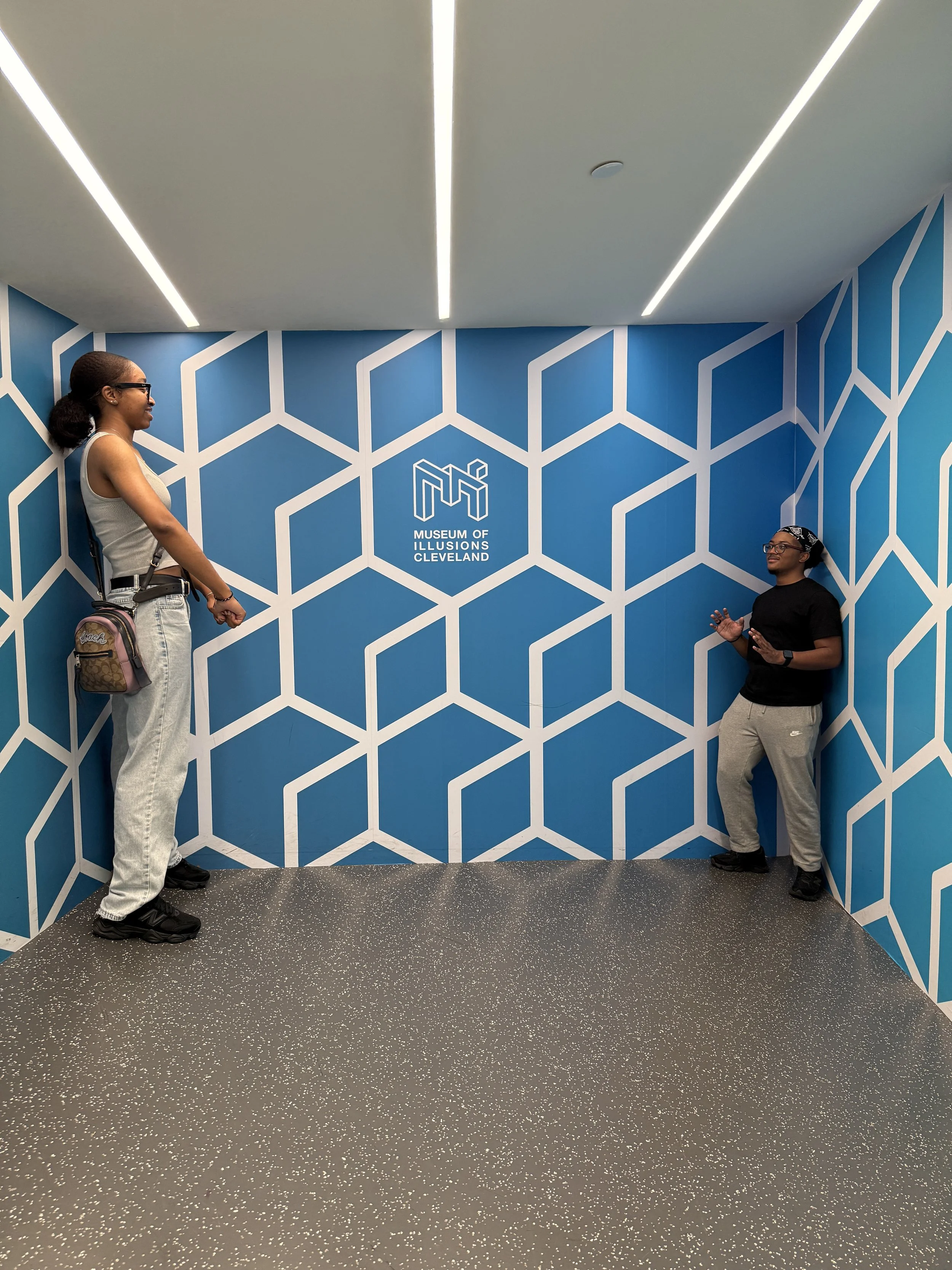 Two women are standing in a corner of the Museum of Illusions Cleveland, which has blue walls with a white geometric pattern and the museum's logo in the center.