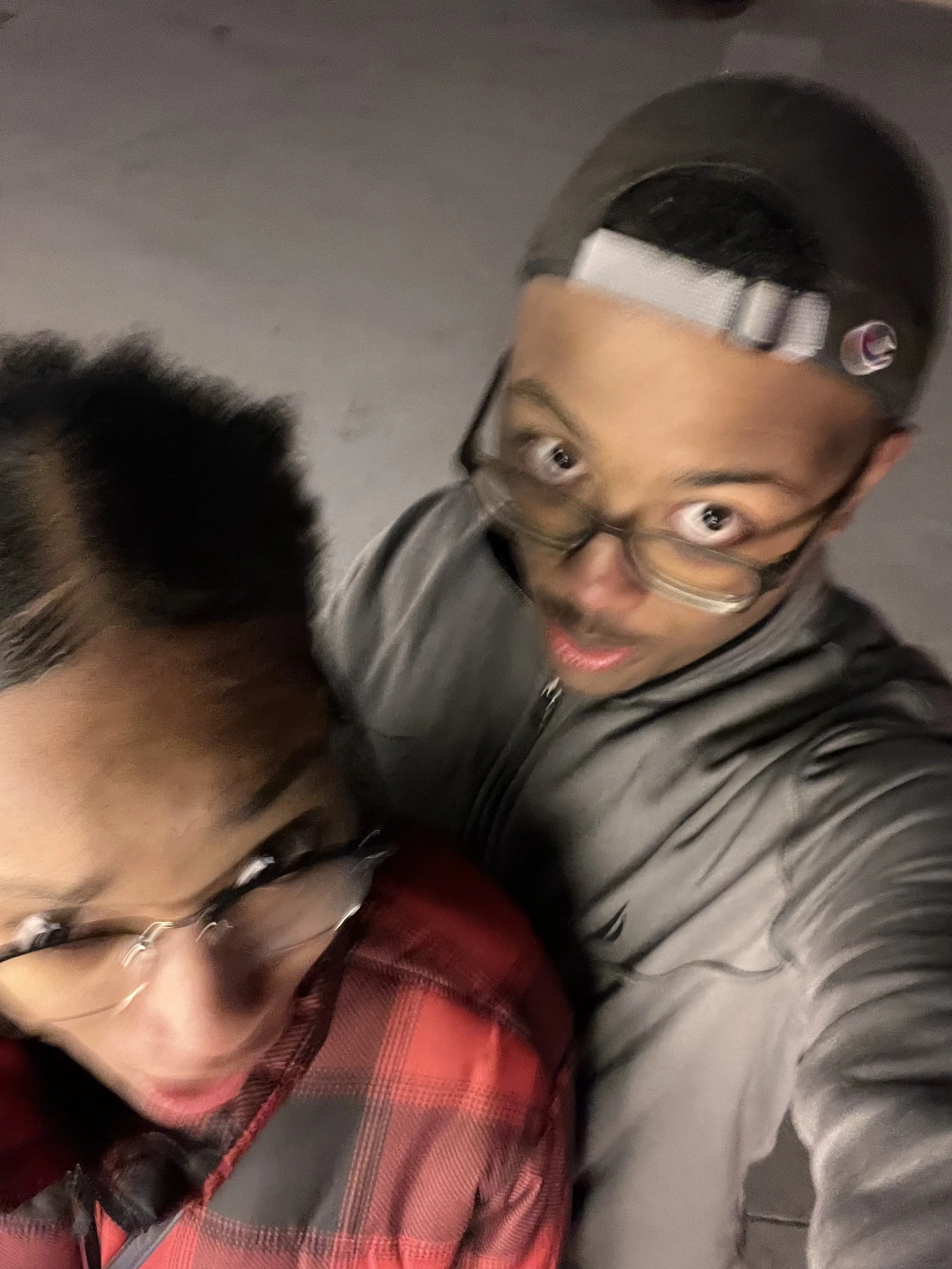 Two young men wearing glasses and jackets taking a selfie from above, with one in a red checkered jacket and the other in a gray jacket and black cap, in an indoor or dark setting.