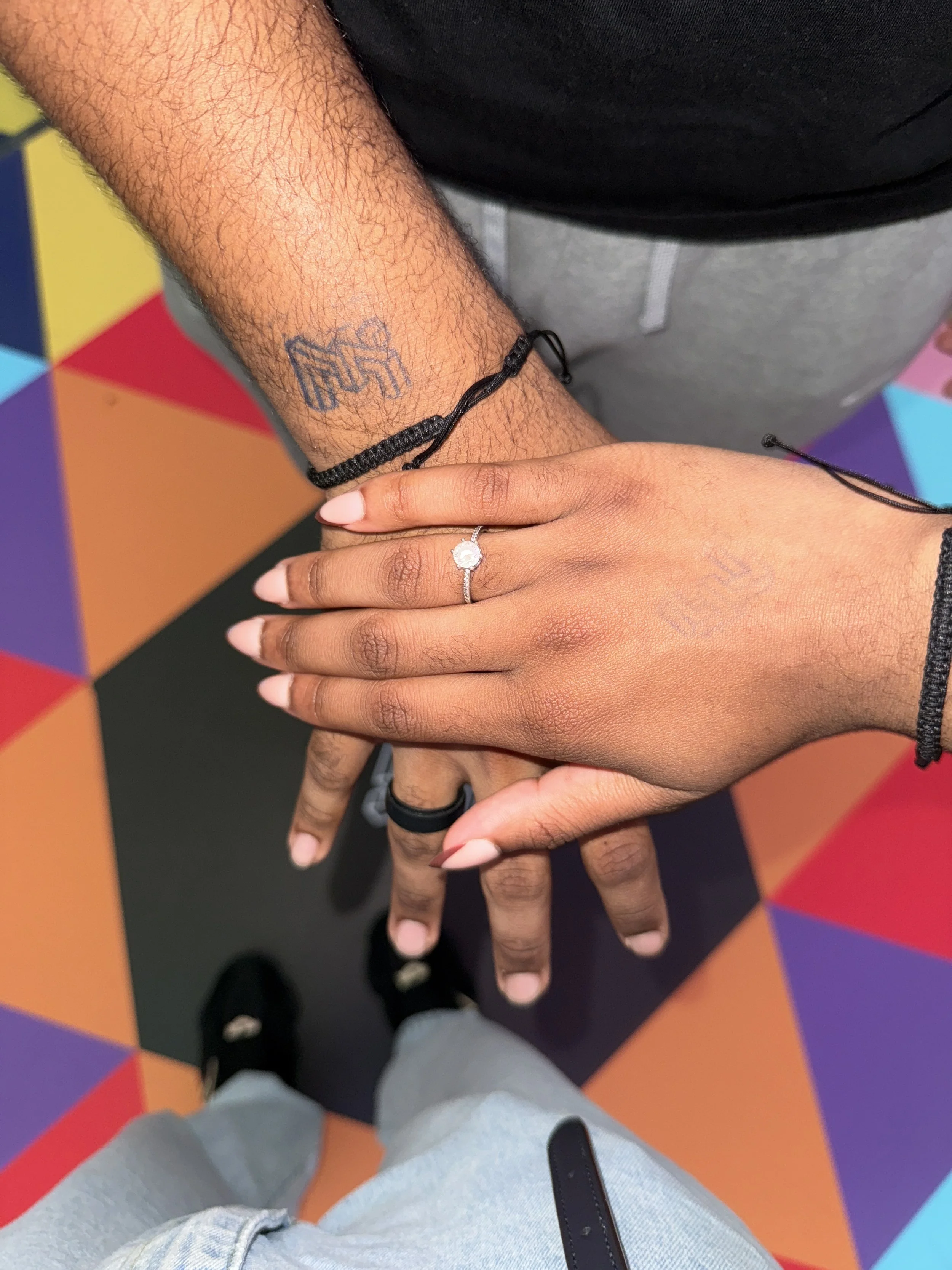 Two hands, one with a ring on the ring finger, are placed together over a colorful tiled floor. The person with the ring is wearing a black bracelet, and the other has a tattoo on the wrist. The photo is taken from a top-down perspective.