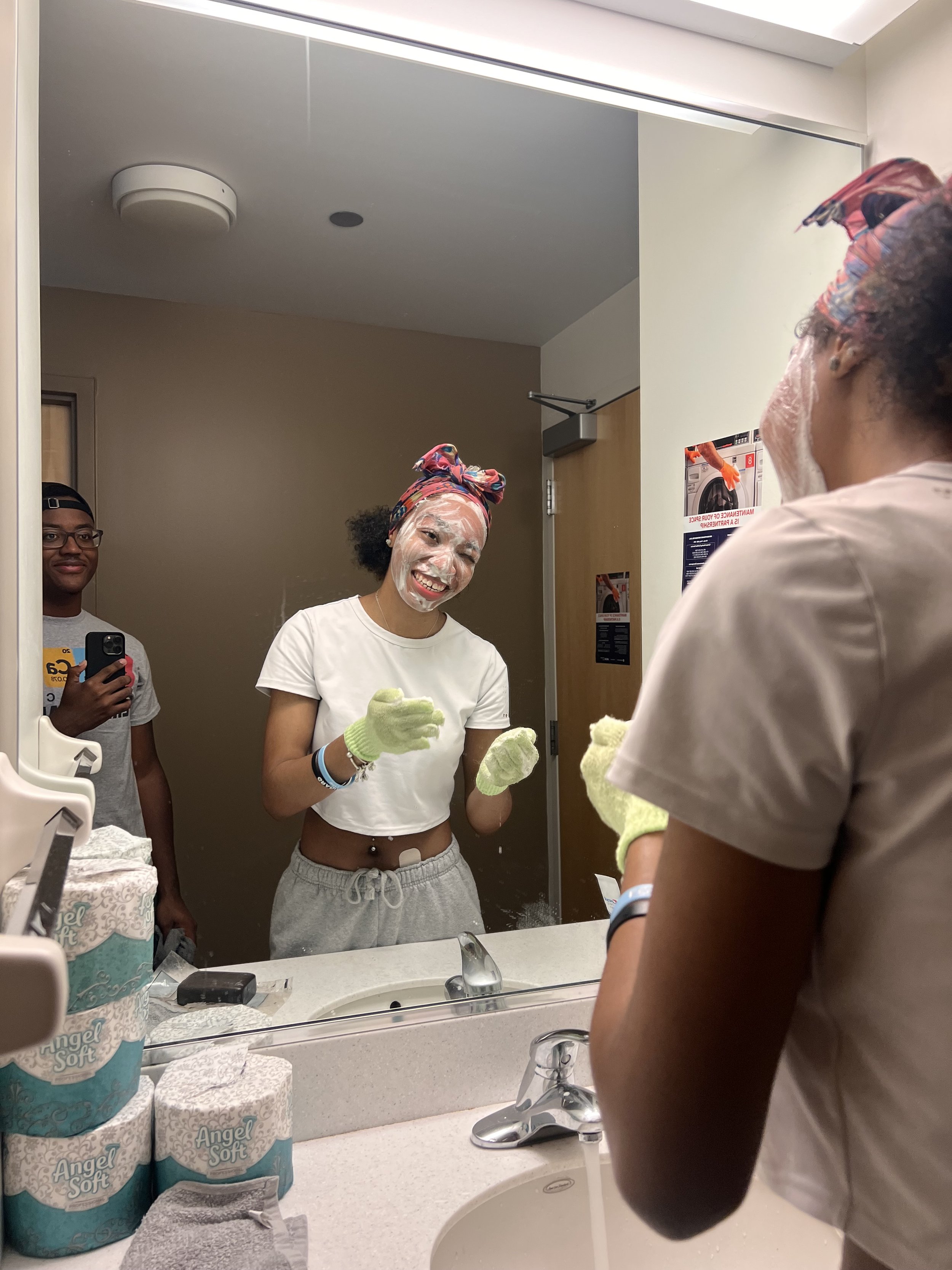 A young woman with soap on her face, wearing green gloves and a colorful head wrap, stands at a bathroom mirror smiling. She is looking at herself while another person, also wearing gloves, is in front of her. A young man is taking a photo of them wi