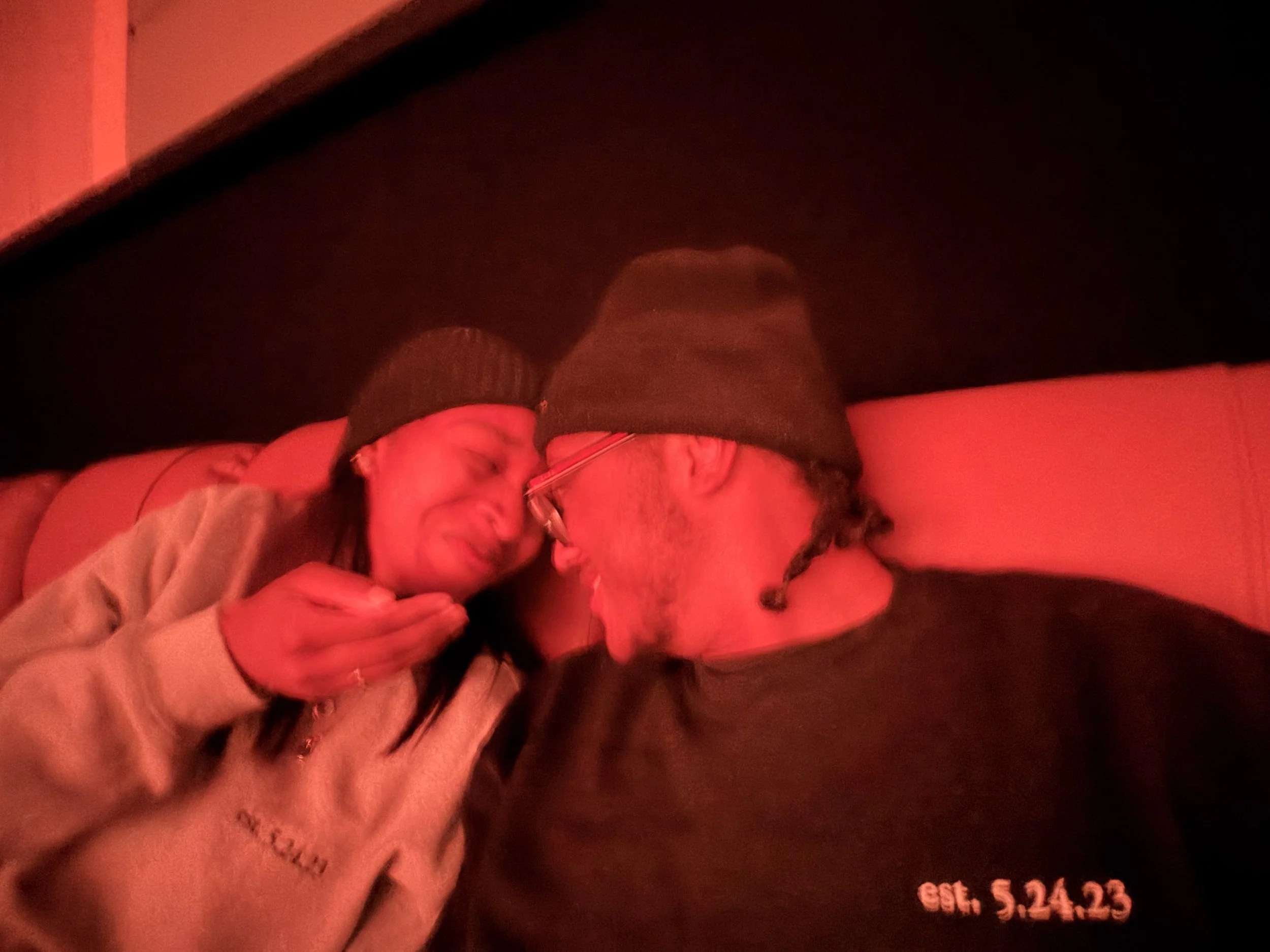 Two people wearing black beanies and glasses, leaning closely together and talking, in a dimly lit environment with red lighting.