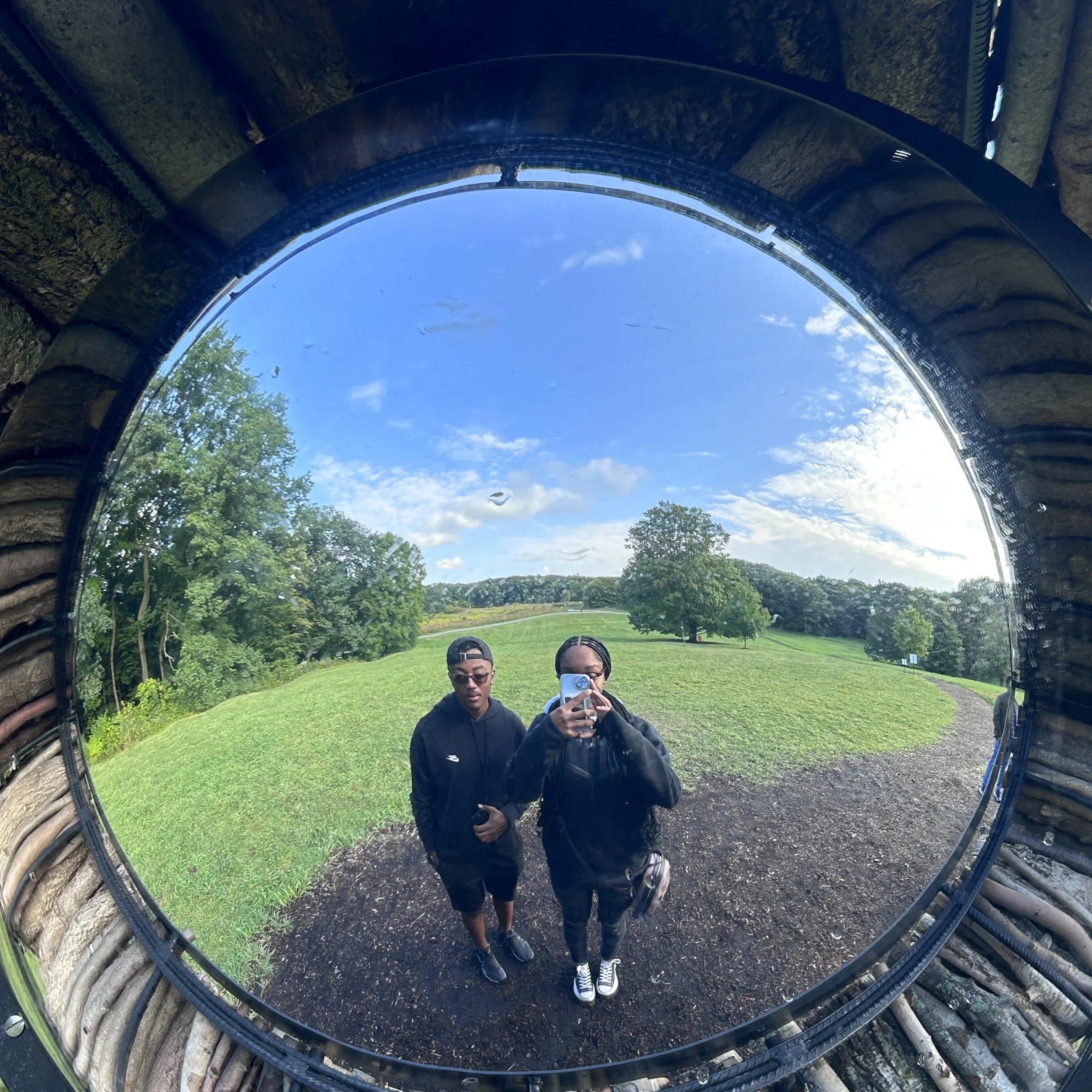 Two young people taking a selfie in a large circular mirror outdoors. The person on the right is holding a phone, and both are dressed in black. Behind them is a grassy hill with trees under a partly cloudy sky.
