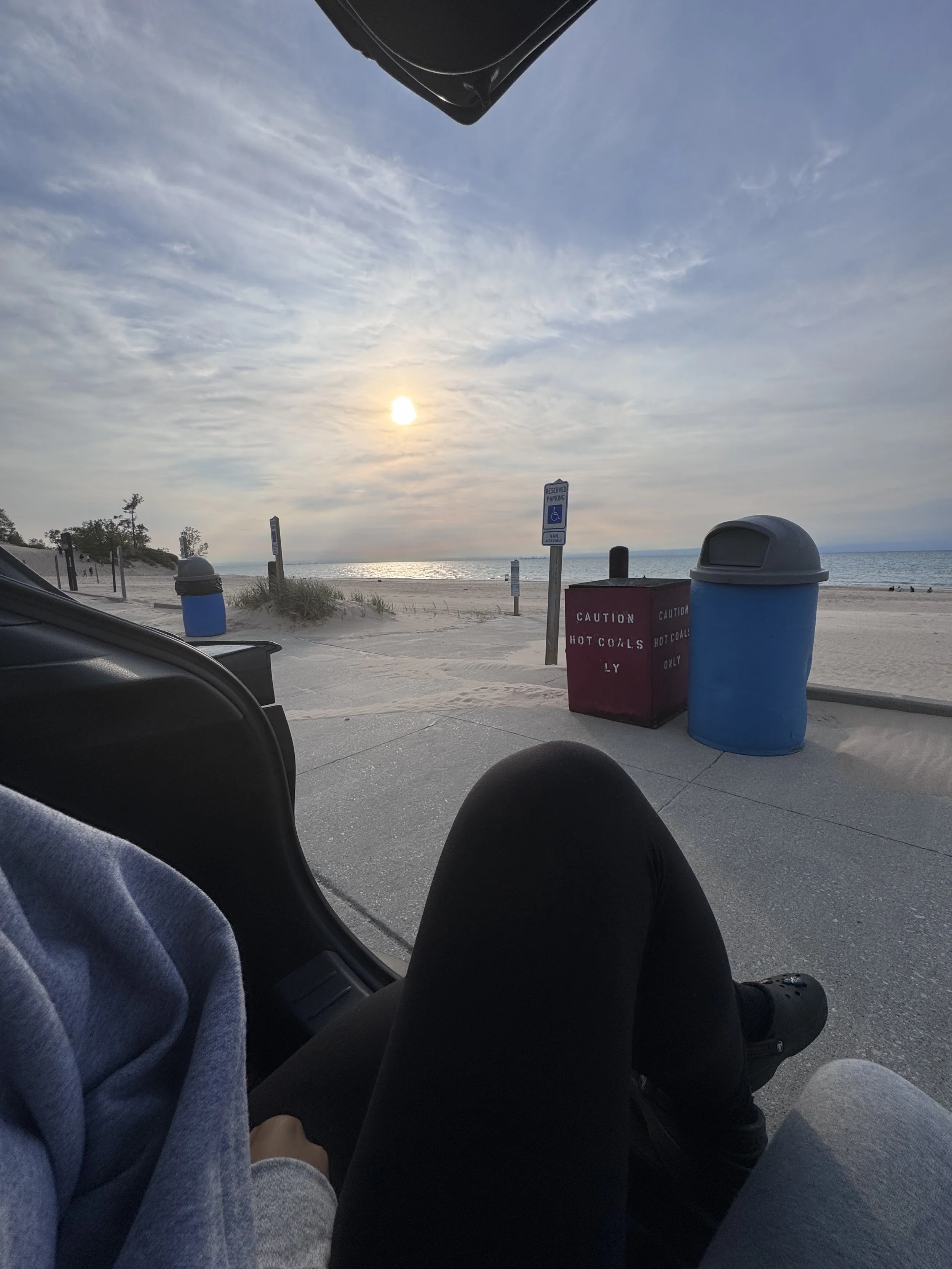 View of a beach at sunset taken from inside a vehicle, with two people sitting inside. The beach has trash bins, a sign for handicapped parking, and a red box labeled 'caution hot coals only'.