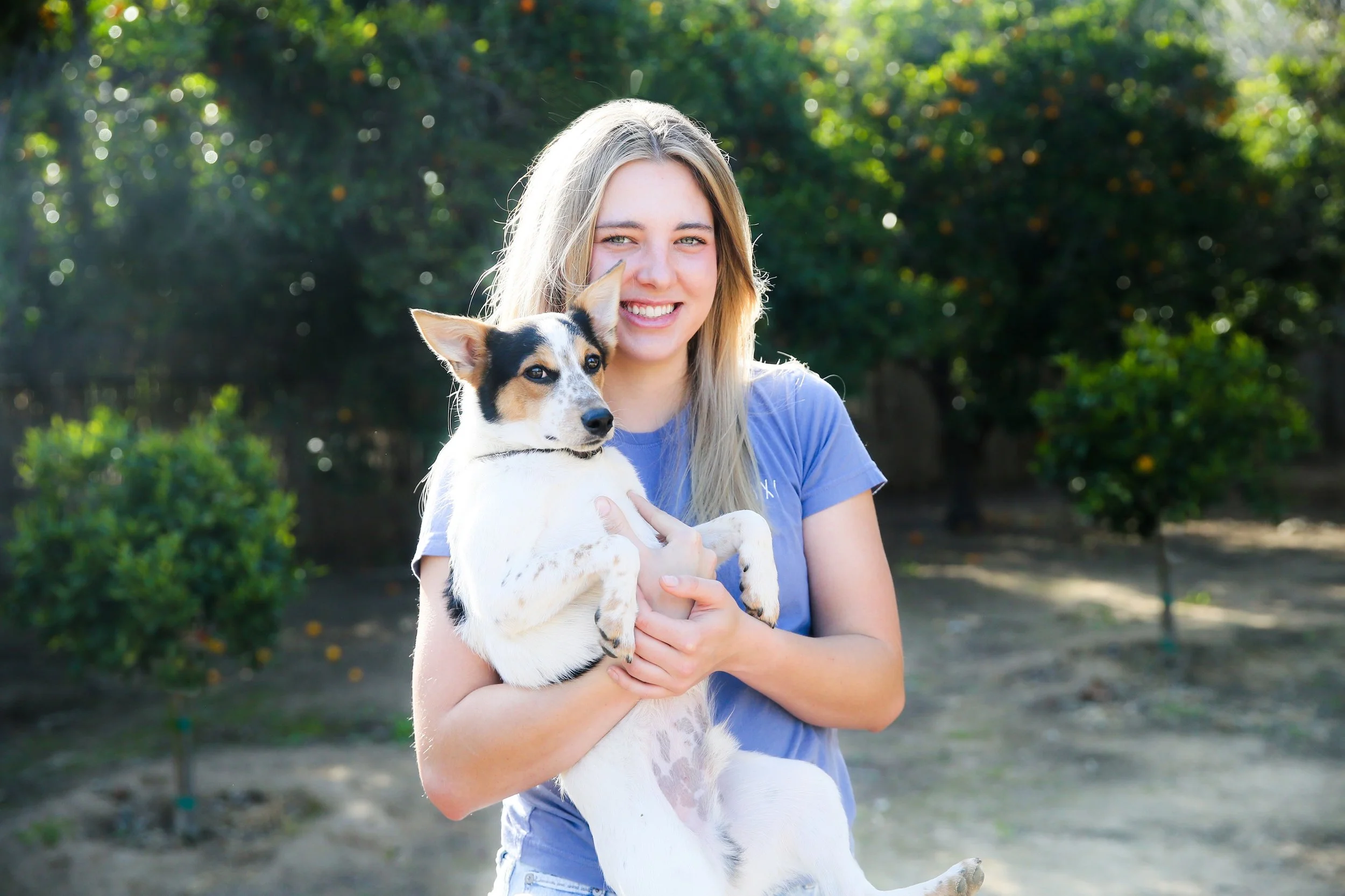 A young woman with blonde hair smiling and holding a small white and black dog outdoors.