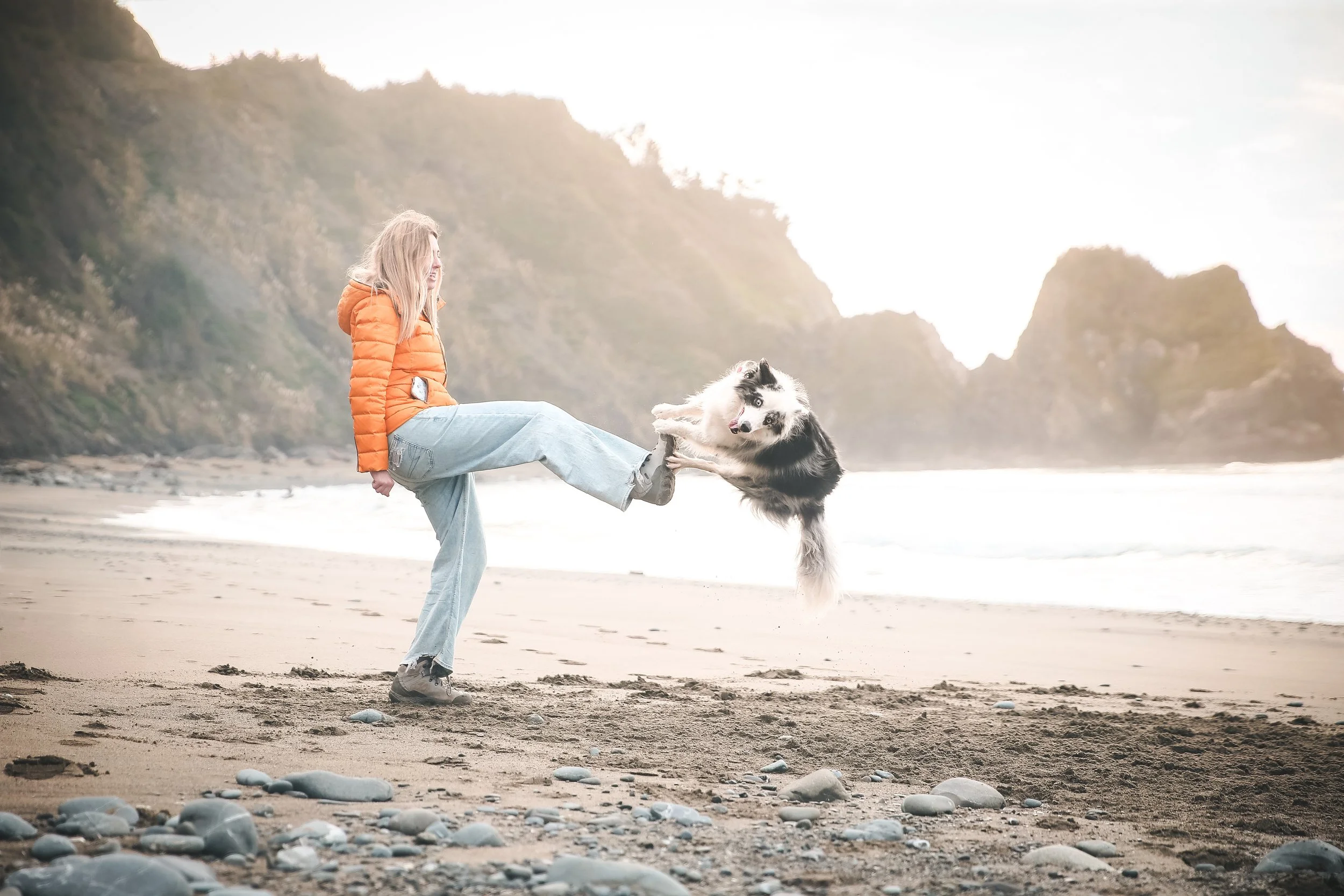 A woman in an orange jacket and jeans kicking a black and white dog on a sandy beach with rocky formations and cliffs in the background.