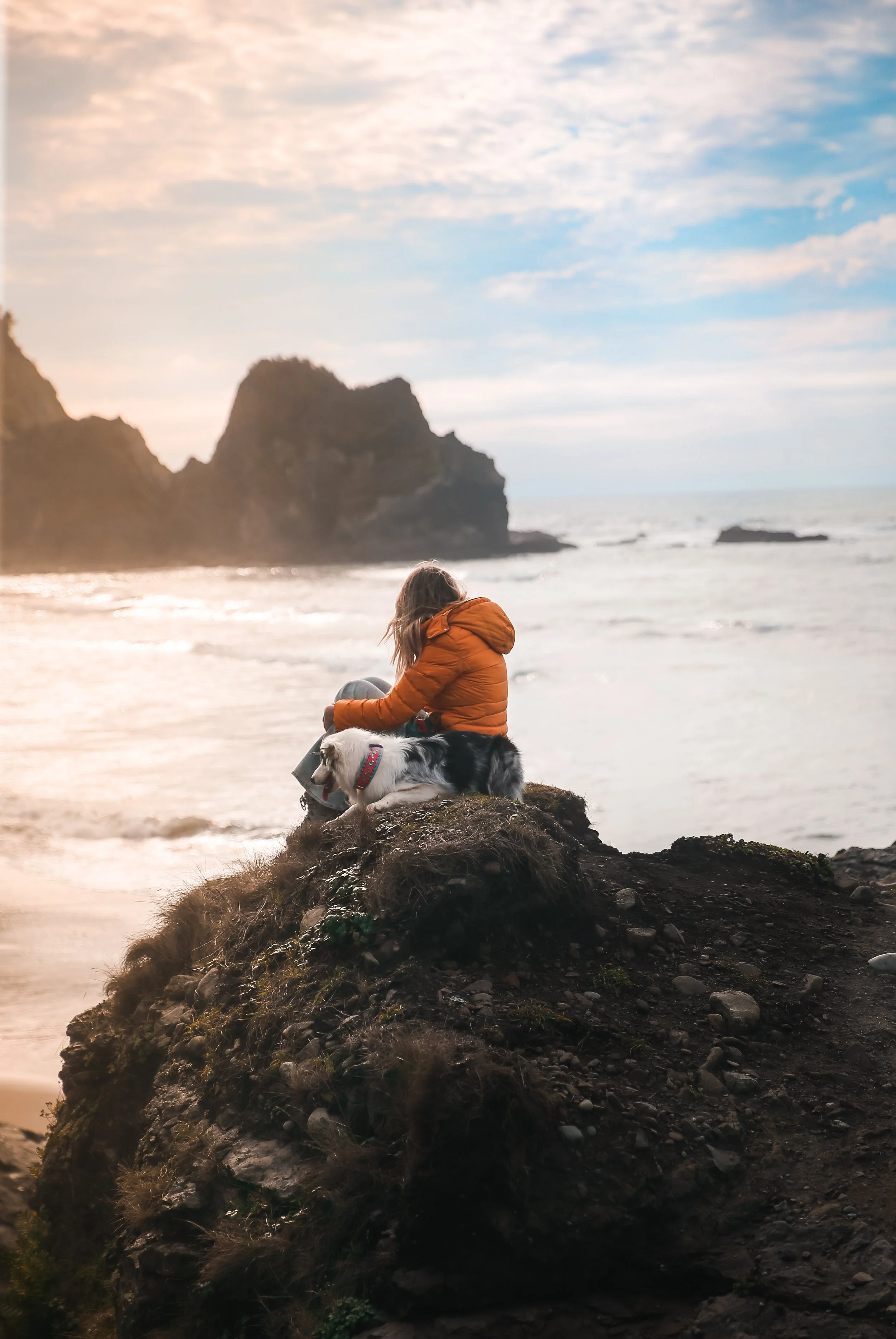 A woman in an orange jacket sitting on a rocky cliff with her dog overlooking the ocean at sunset or sunrise.