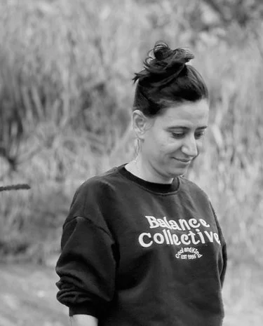 A woman with a bun hairstyle wearing a black sweatshirt with the words 'Balance Collective' in white, standing outdoors with tall grasses in the background.