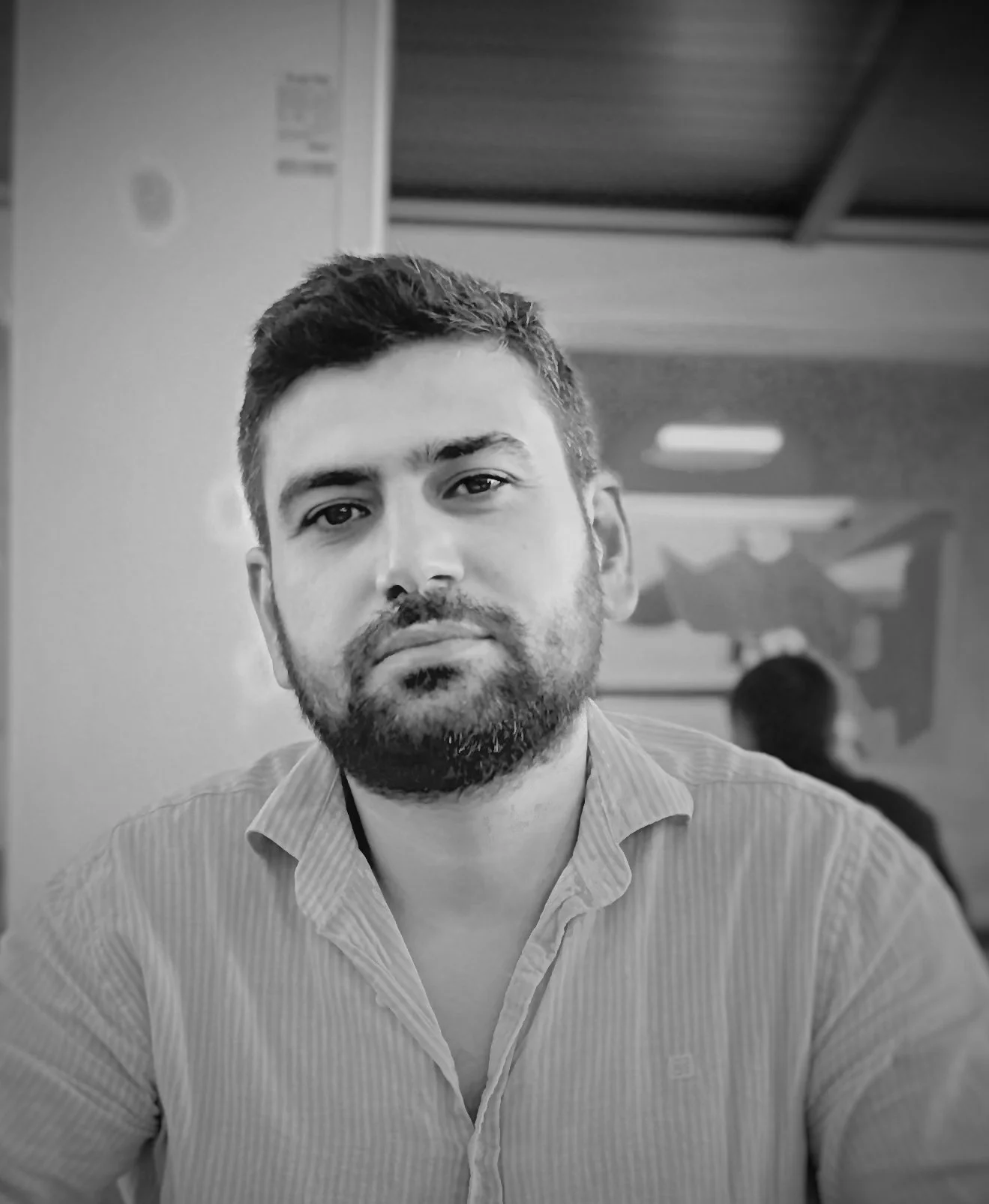 Black and white photo of a man with short hair and a beard, wearing a collared shirt, sitting indoors with a blurred background that includes another person and wall art.