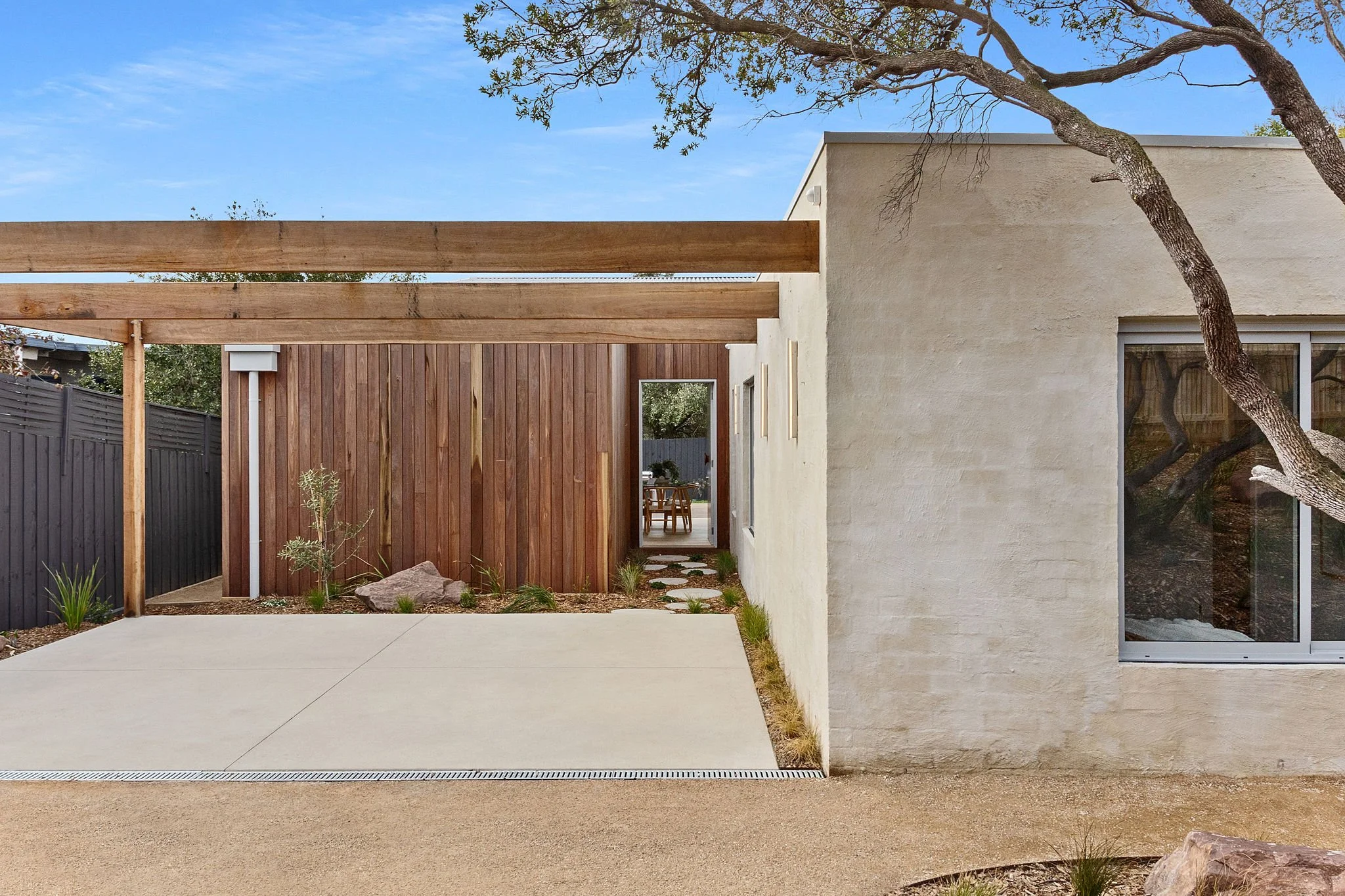 Modern house exterior with a concrete patio, wooden accents, a tree, and a window with a curtain inside.