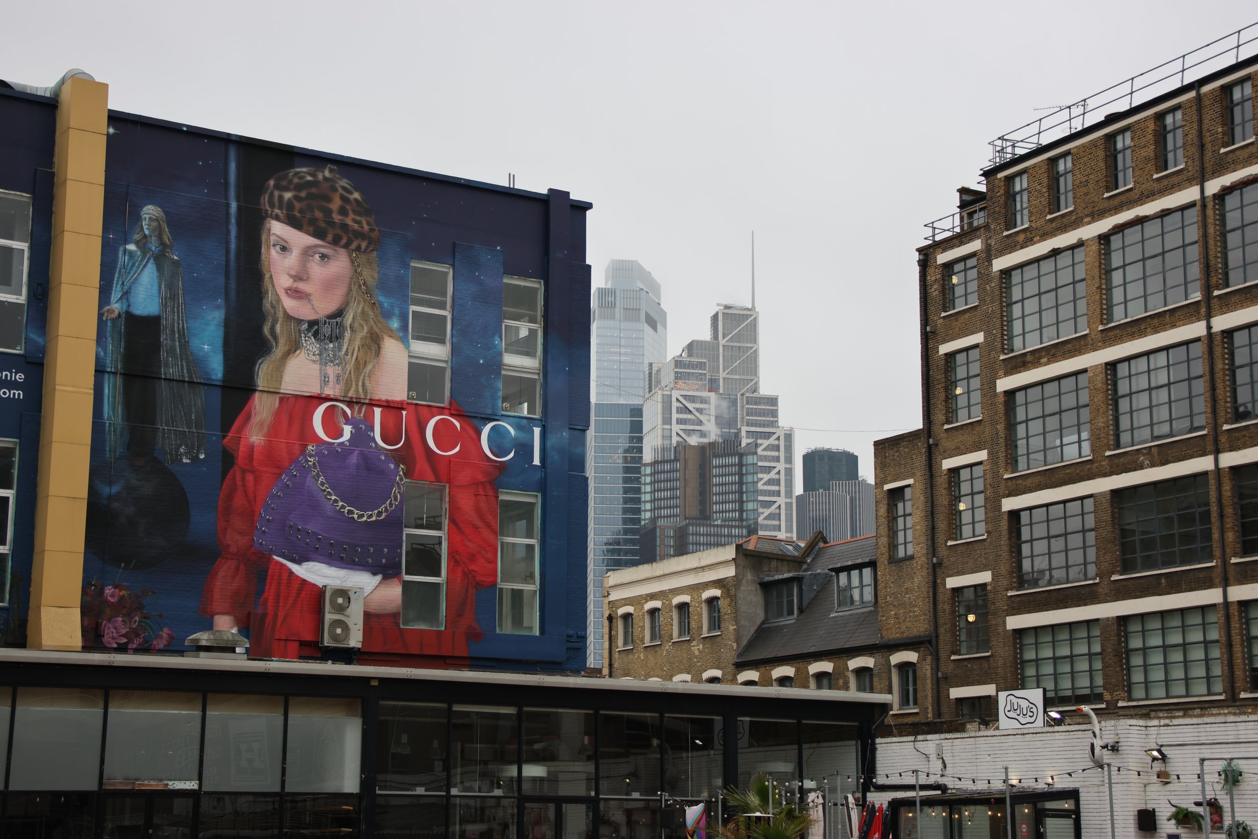 Large Gucci billboard featuring a girl with blonde hair wearing a leopard print cap, red jacket, and choker, with city skyscrapers in the background.