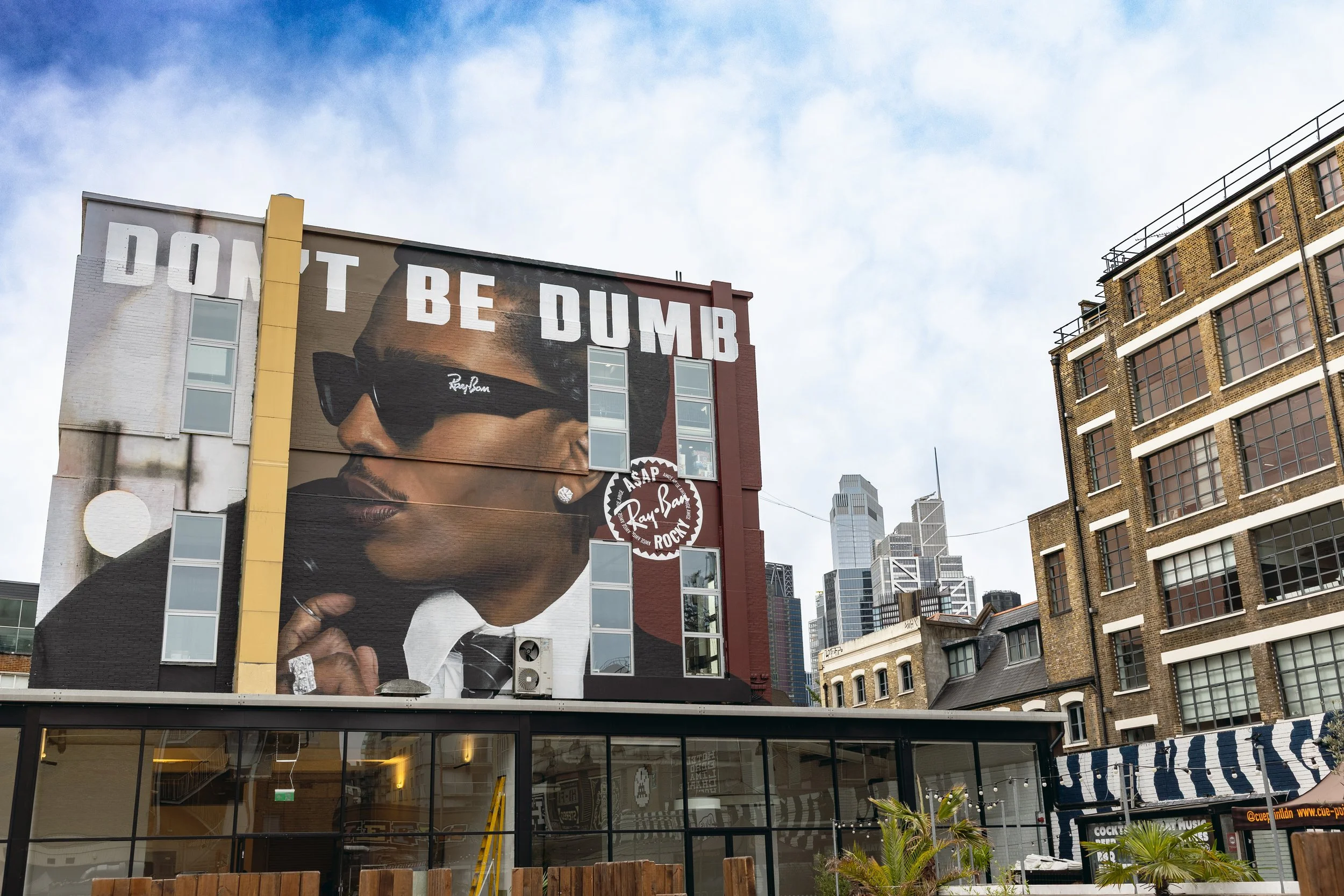 Large billboard on building featuring a portrait of a woman with sunglasses and earrings, with the text 'DON'T BE DUMB' at the top.