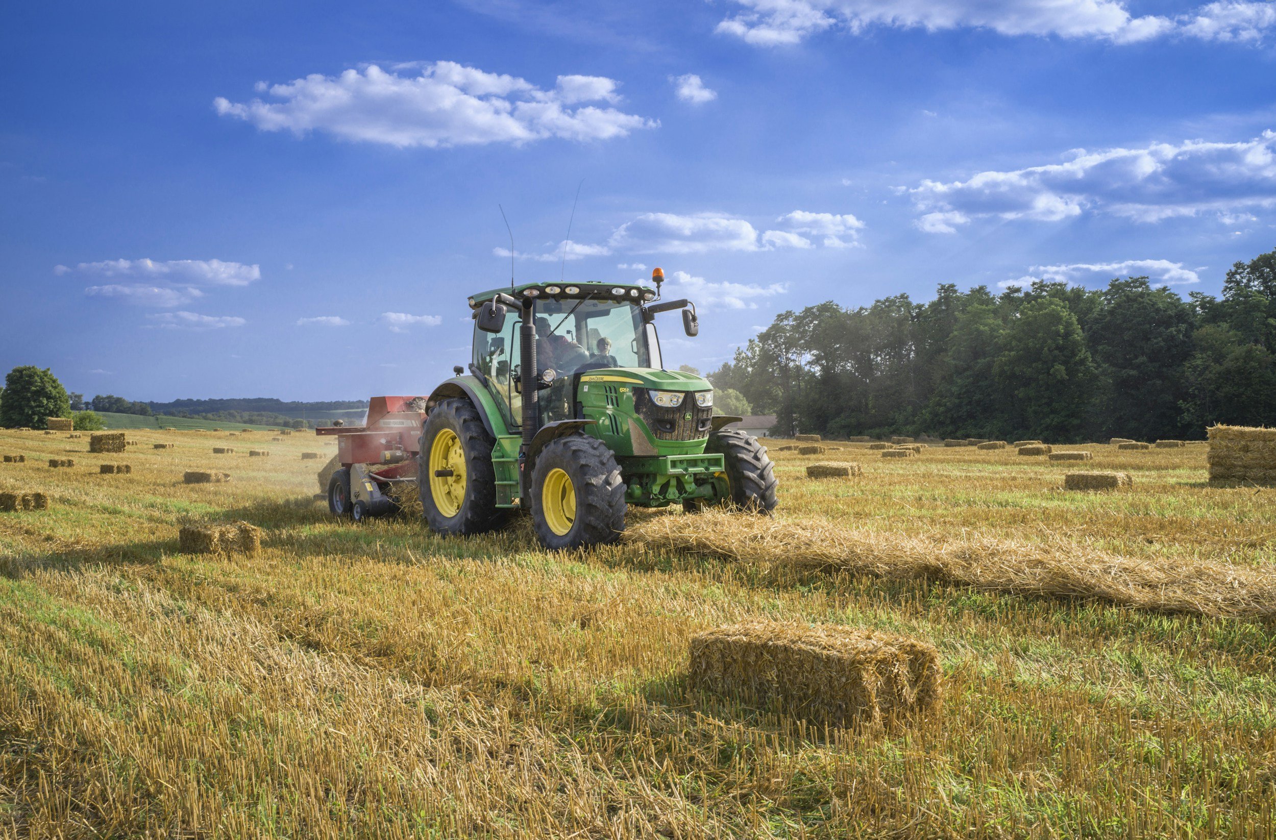 A green tractor with yellow wheels is working on a hay field, pulling a hay rake behind it. The field has hay bales scattered across it, with a line of trees in the background under a blue sky with scattered clouds.