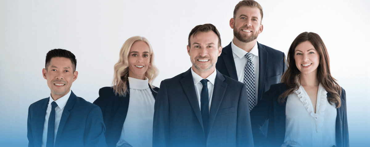 Group of five diverse business professionals in formal attire smiling in an office setting.