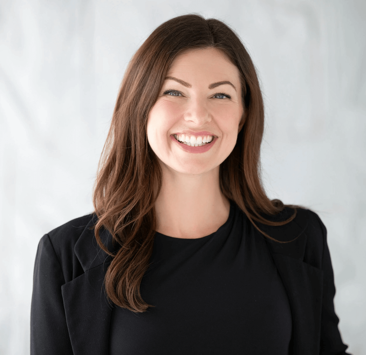 A woman with long brown hair wearing a black top and blazer, smiling against a plain light background.