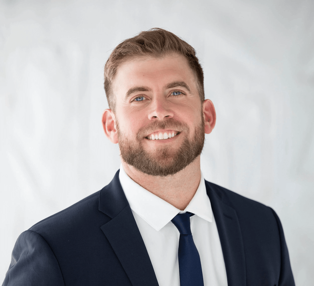 A man with a beard and short brown hair, wearing a navy suit, white shirt, and navy tie, smiling against a light gray background.
